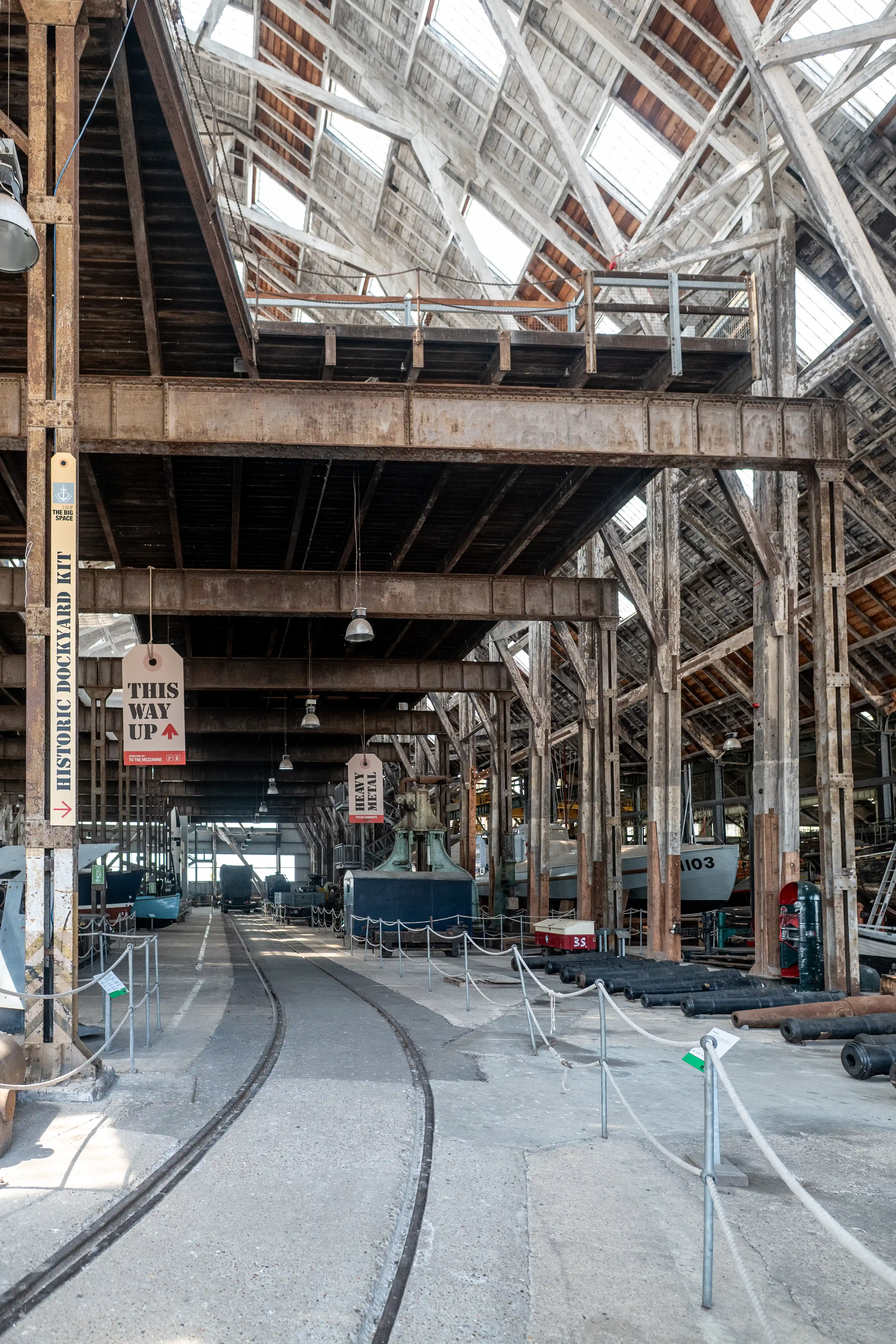 Interior view of a large, old dockyard building with exposed wooden beams, metal gantries, and a glass-panelled roof. In the foreground, a curved railway track runs past displays of cannons and naval artefacts, including a boat labelled 1103. Informational signs are visible.