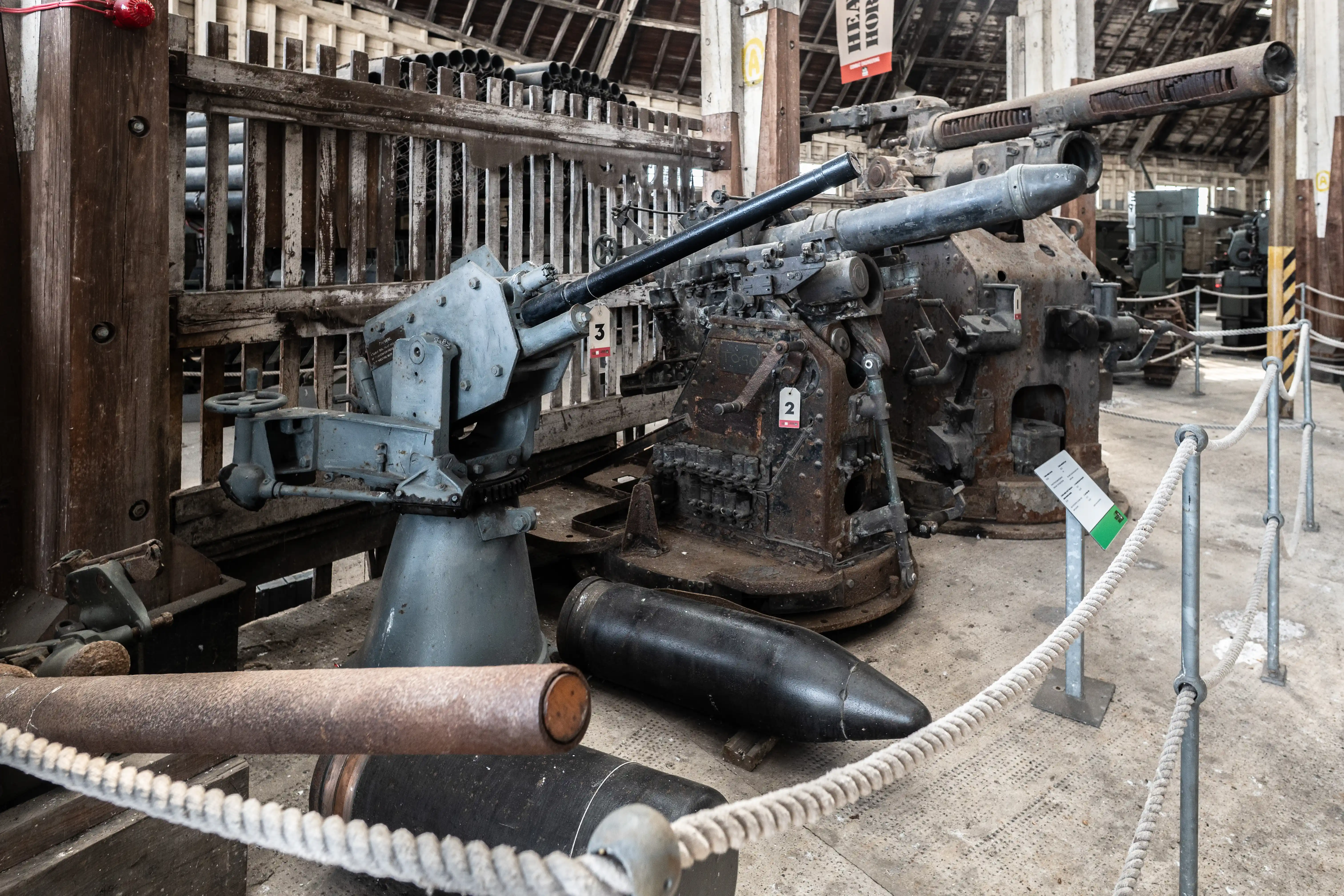 A museum display of antique artillery pieces and large shells, with focus on a grey anti-aircraft gun and a rusted, larger cannon.