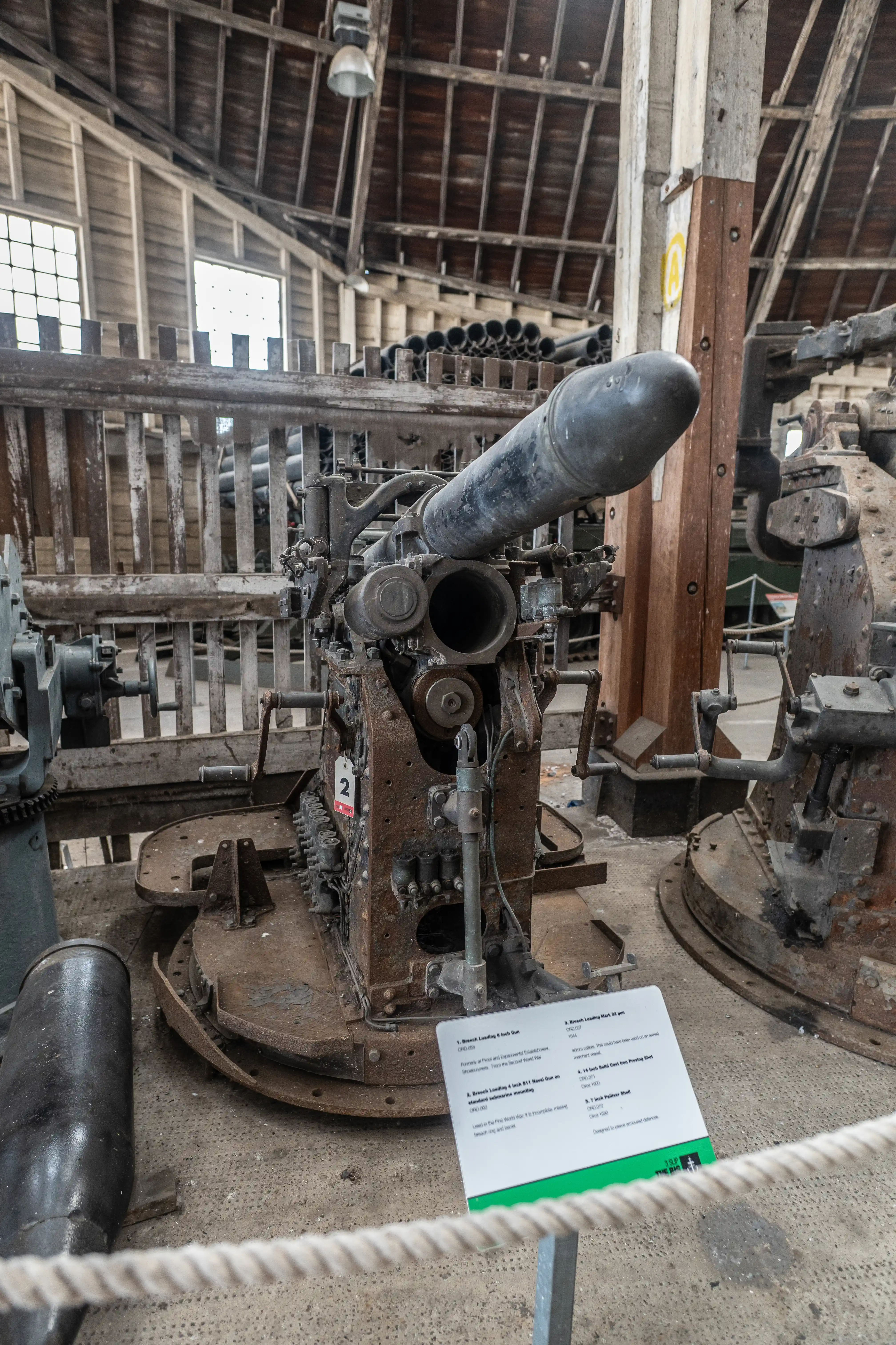 A heavy, dark grey naval gun with a thick barrel and complex machinery is displayed in a barn-like building, with a shell lying on the floor to its left.