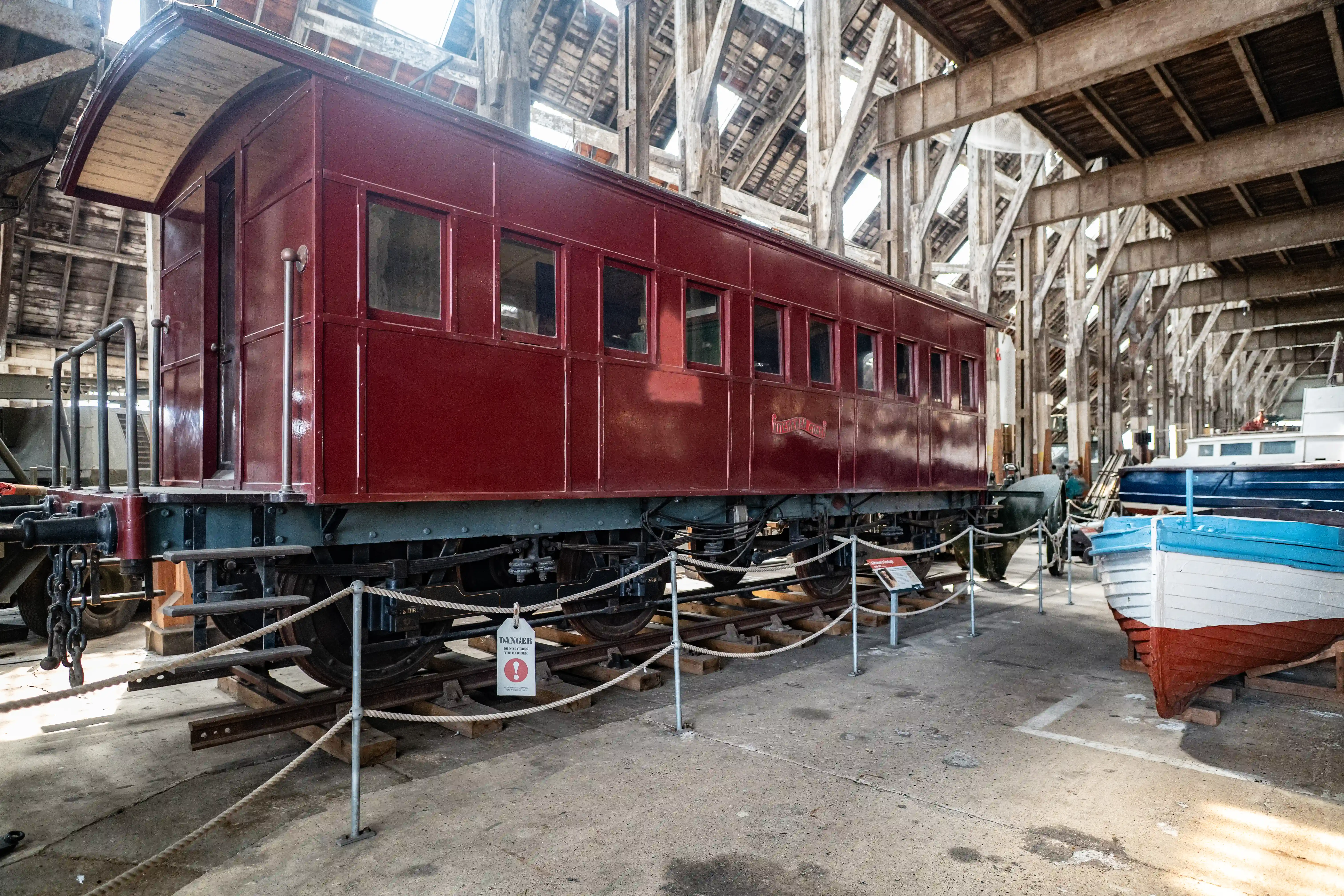 A detailed view of the side of a vintage maroon railway carriage with multiple windows, resting on railway tracks inside a cavernous warehouse with exposed wooden beams. Several boats are visible in the background.