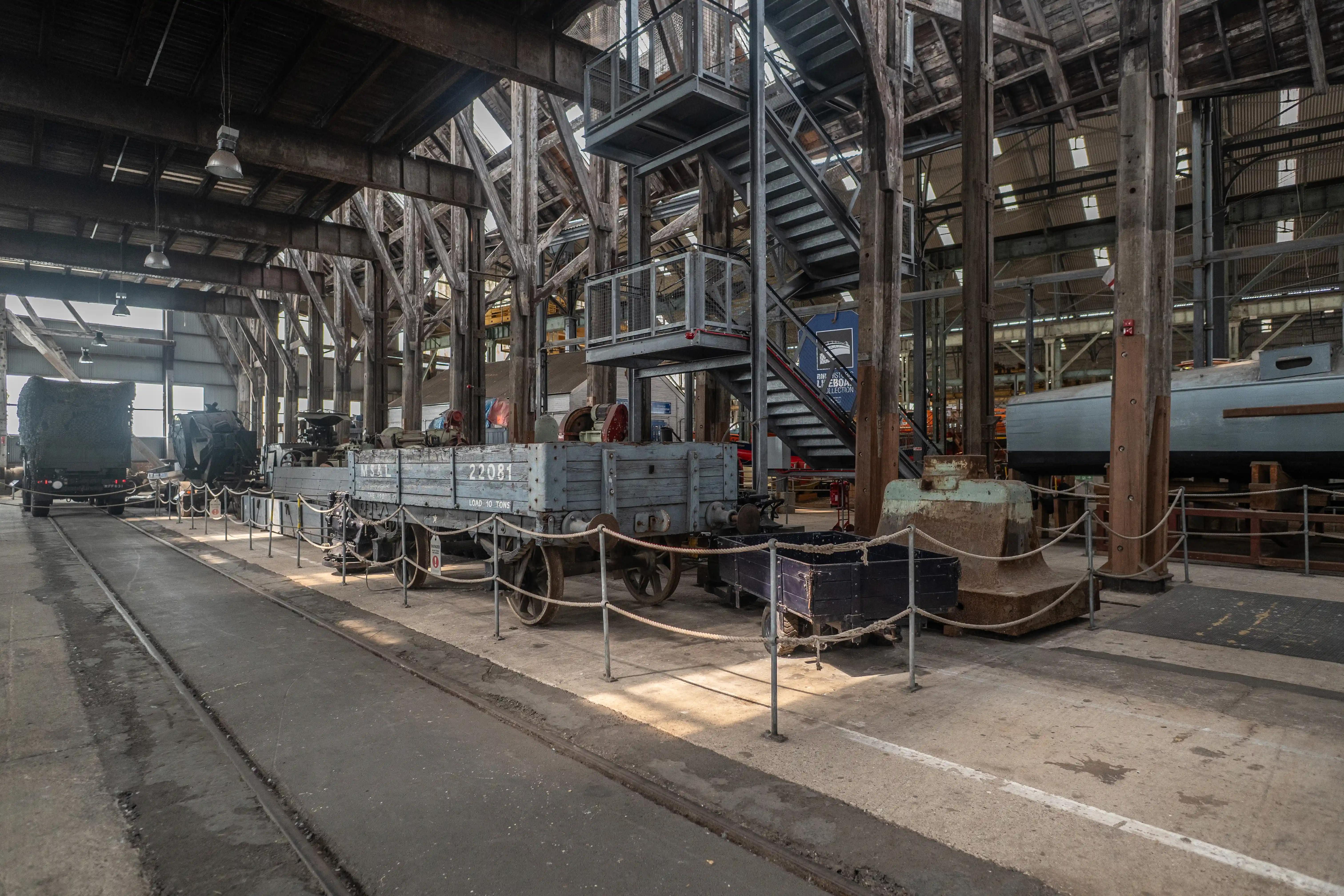  Interior view of a grand, old railway workshop with wooden and metal beams, vintage grey wagons on tracks, a large industrial machine, and a grey boat on stands.