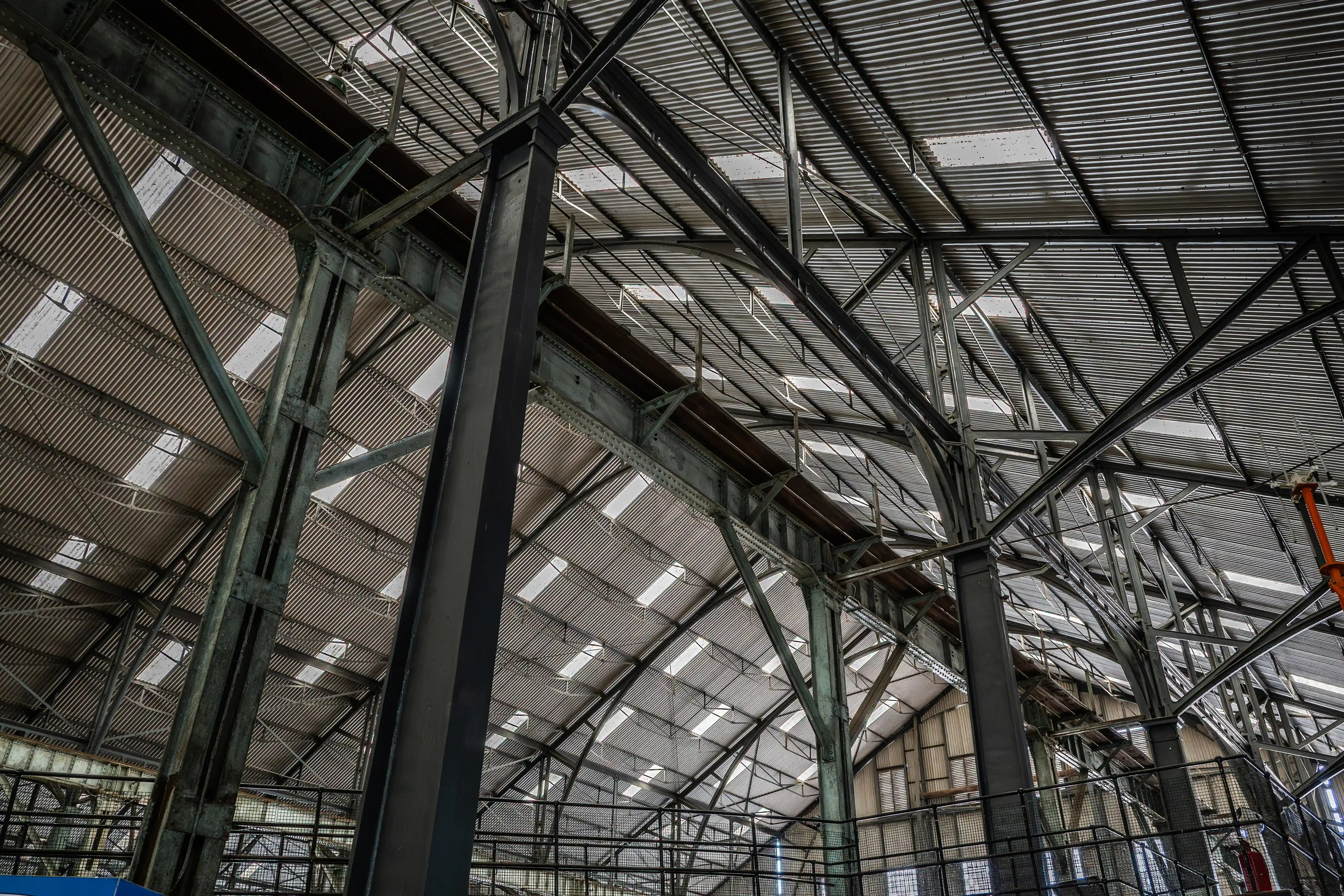 A low-angle shot of the underside of a large industrial building's roof, showing a complex network of dark grey steel beams, columns, and trusses supporting corrugated metal roofing with rectangular skylights.