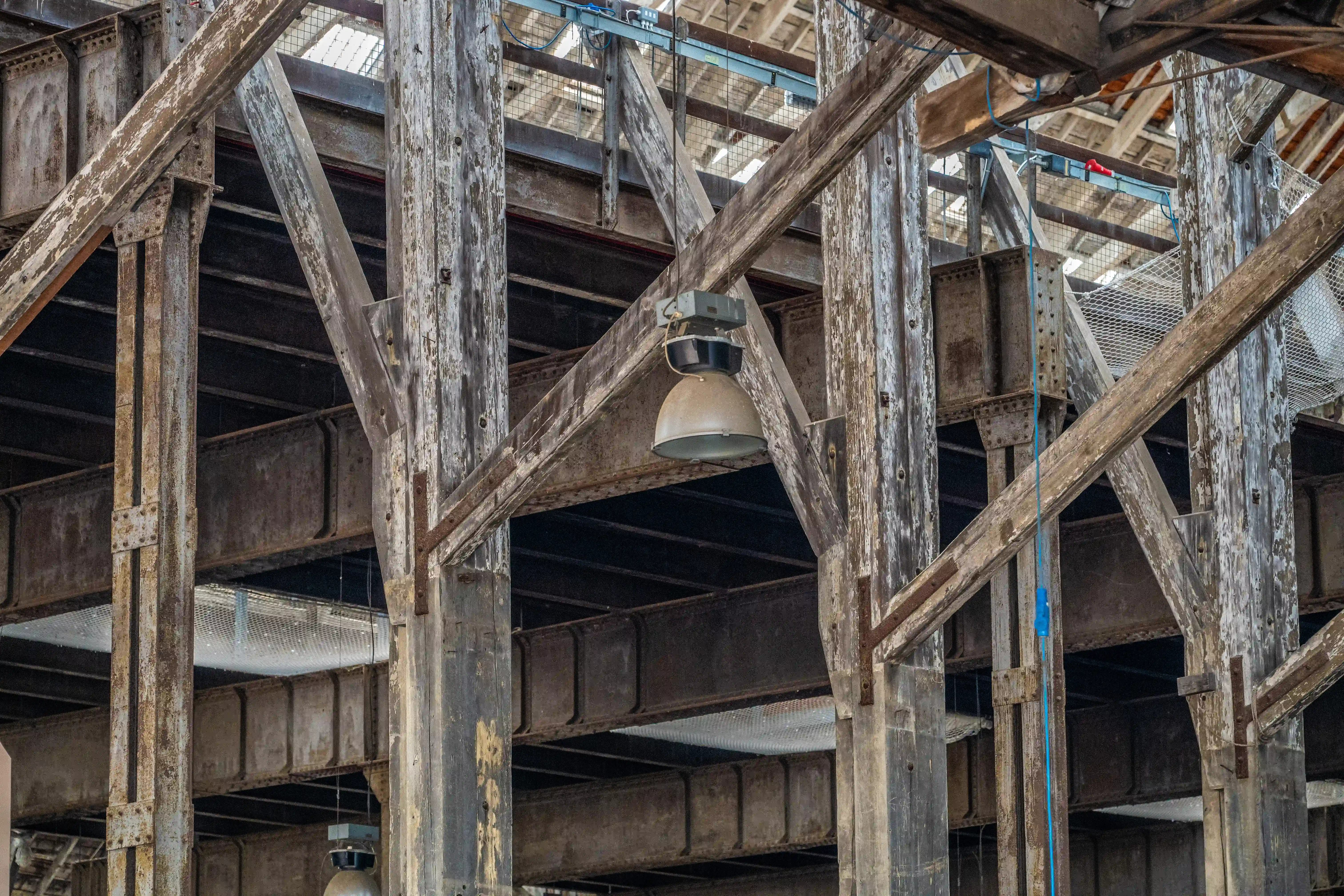  Interior of an industrial building with weathered wooden posts and beams crisscrossing with exposed steel girders. A large bell-shaped industrial light fixture hangs from above.