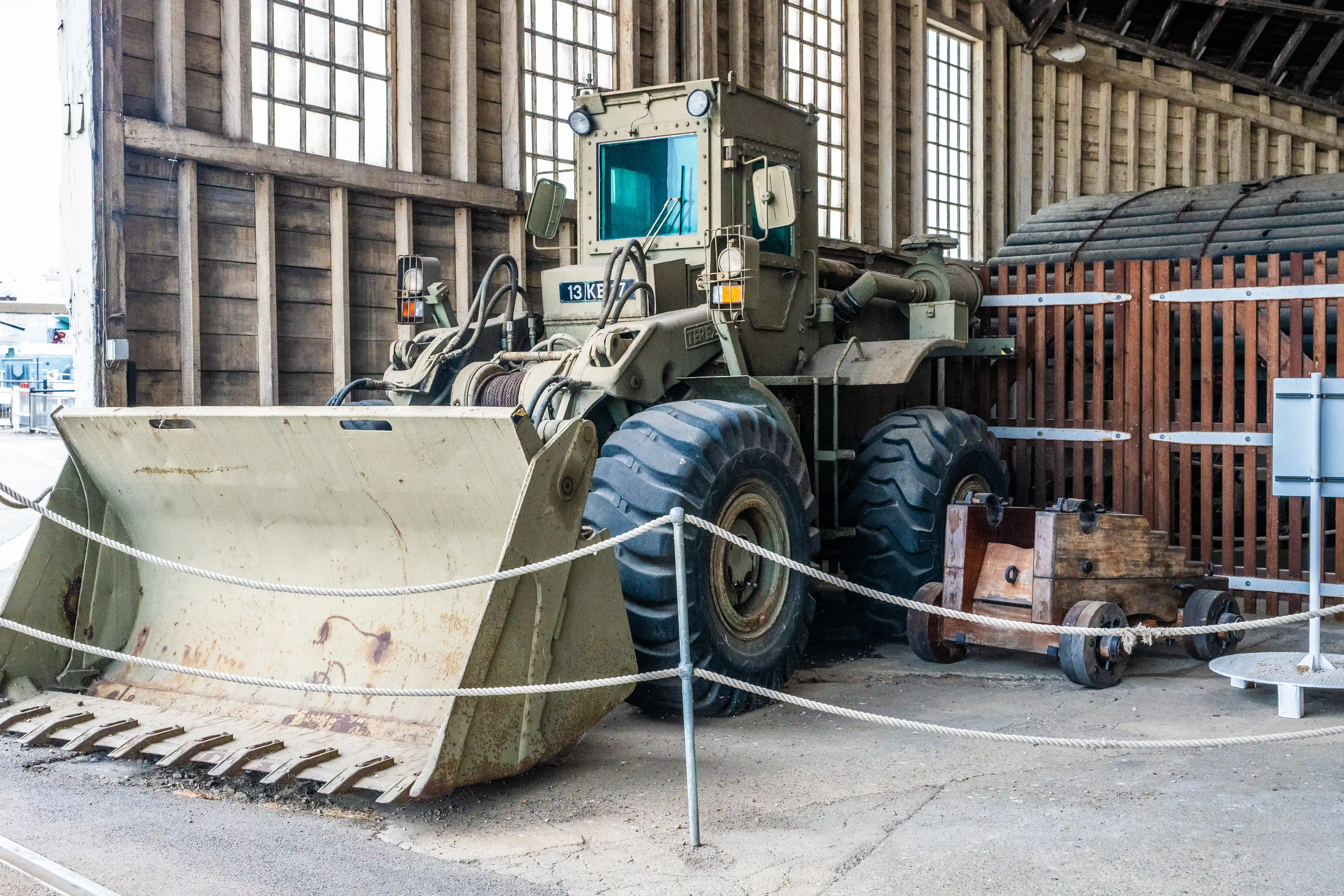 A large, olive-green earthmover with a wide bucket and large tyres is parked indoors, with a small wooden cart to its right.