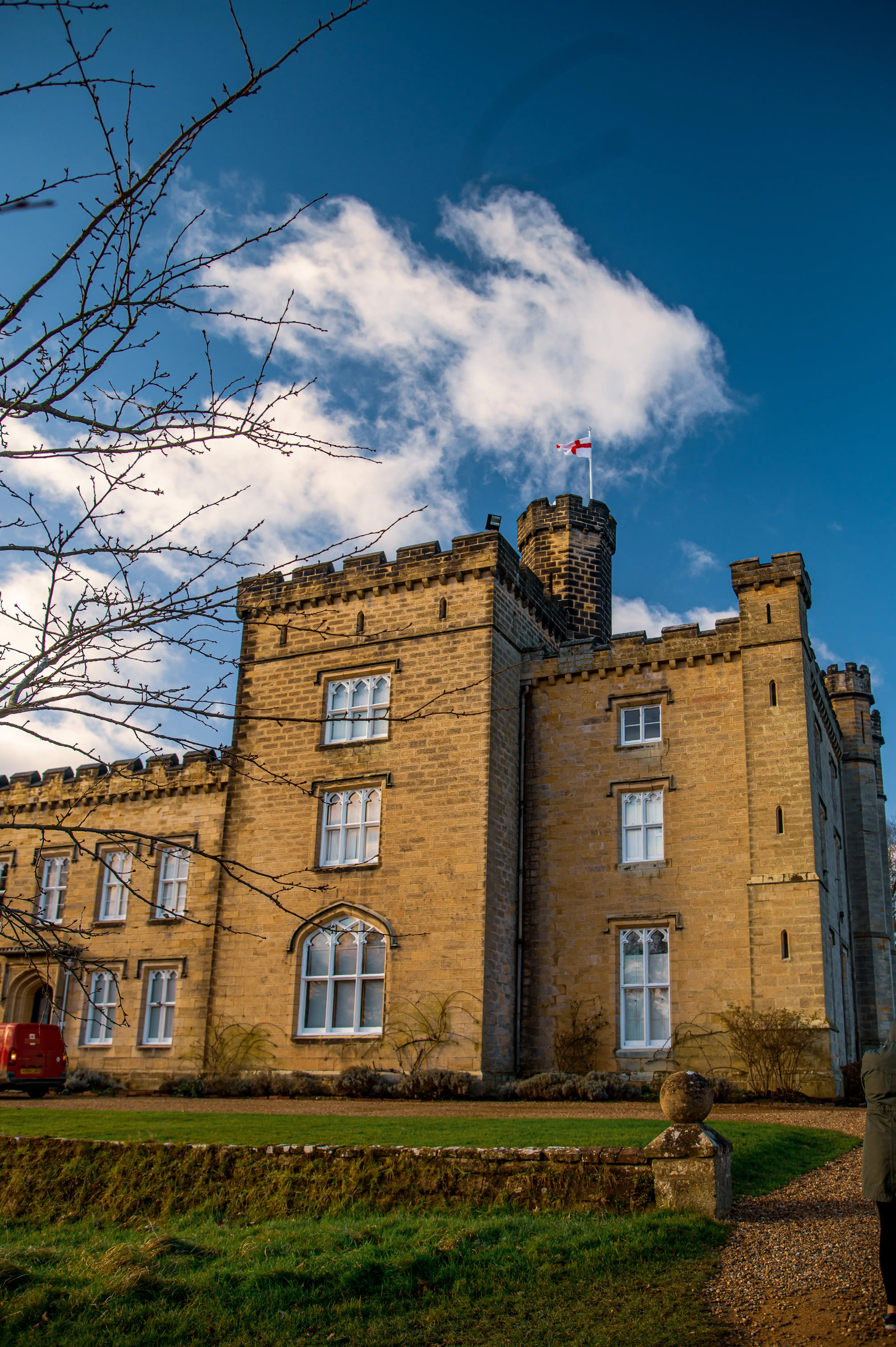 A large, imposing castle constructed of yellow brick, featuring crenellated towers and arched windows with white frames. The building is set against a bright blue sky with scattered white clouds. A bare tree branches reach into the frame from the left. An English flag is visible atop one of the turrets.