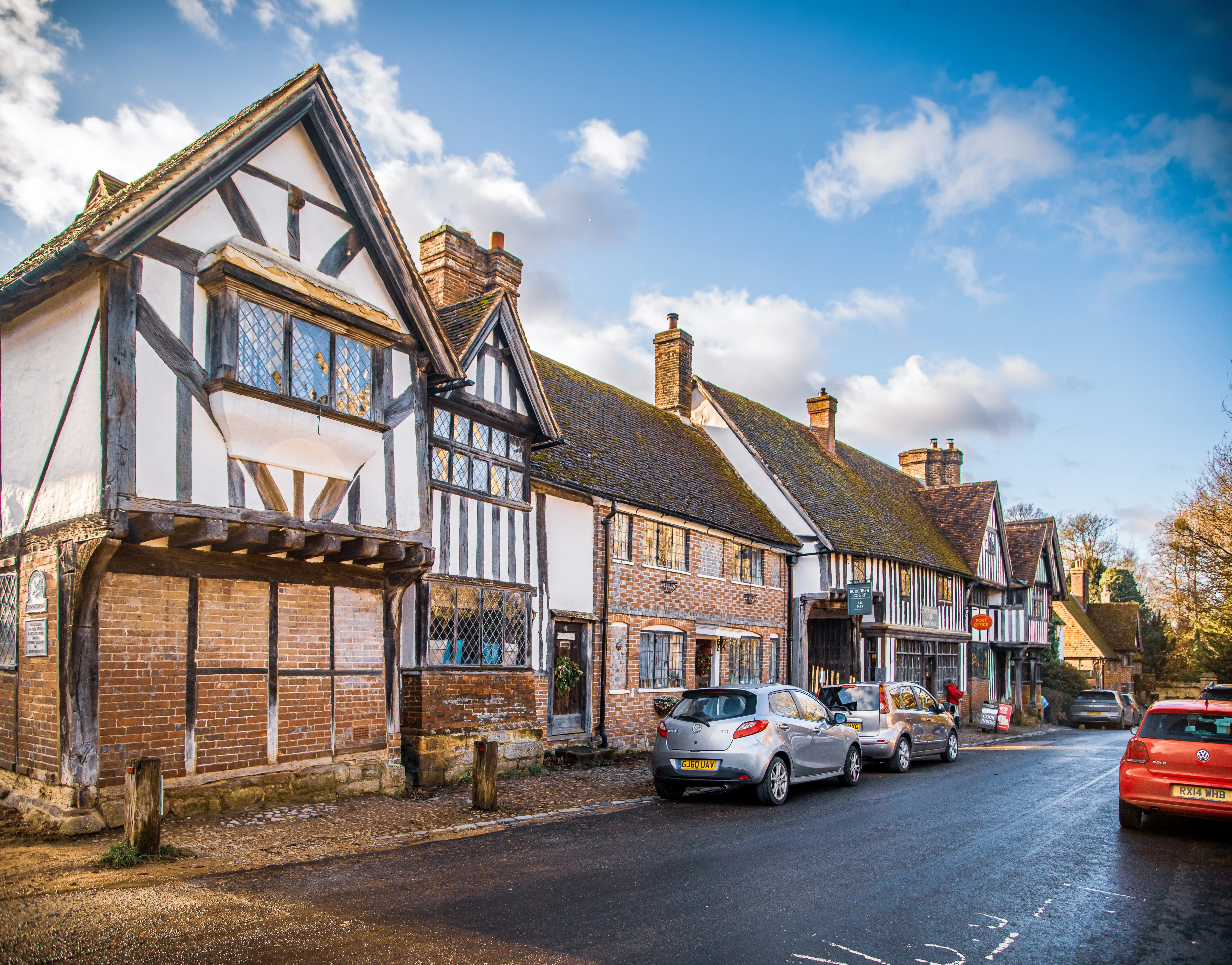 A row of historic buildings with timber framing and leaded windows on a street in an English village, with cars parked and a blue sky with clouds.