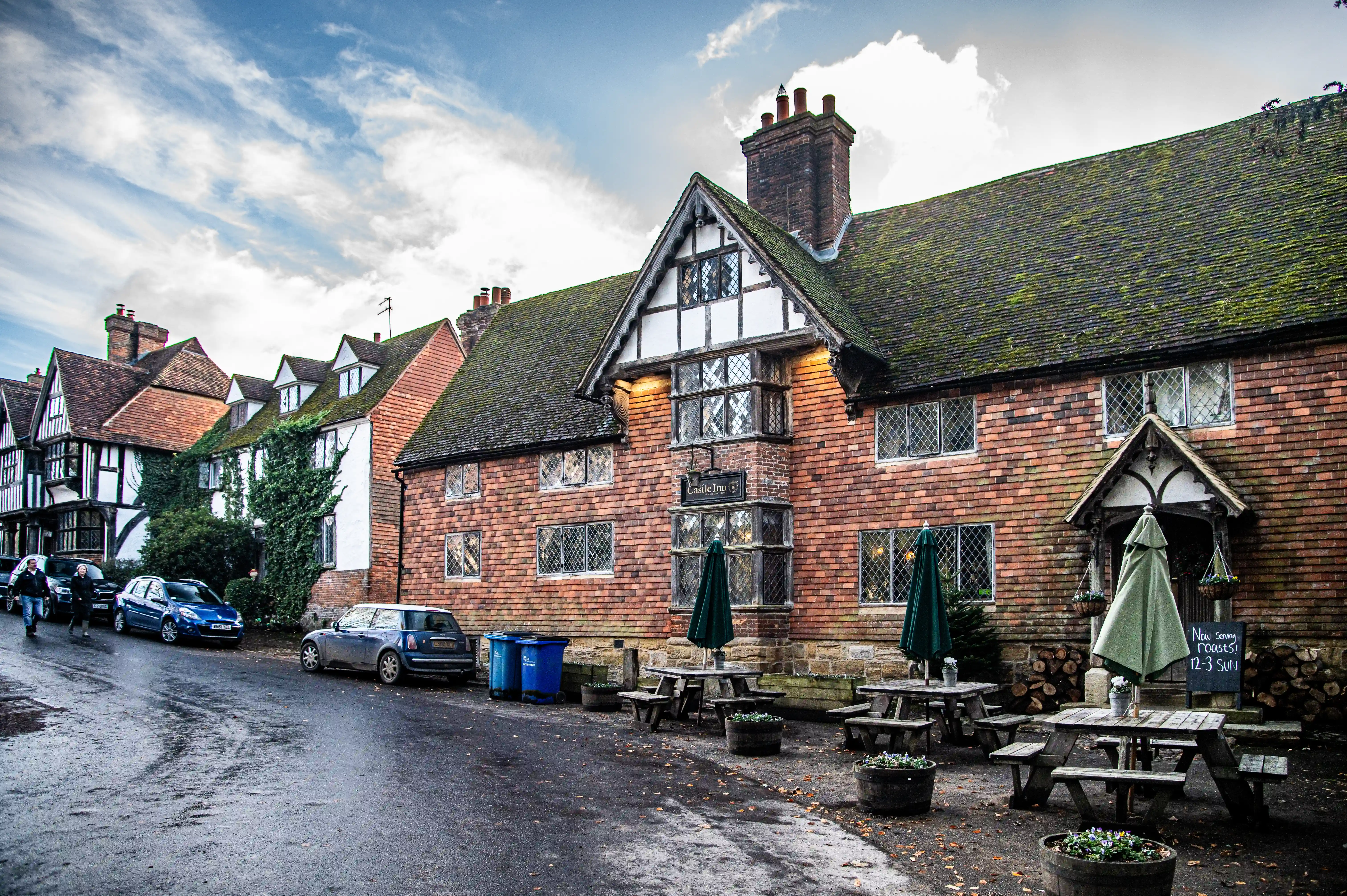 Exterior view of the Castle Inn, a historic pub with red brick walls, timber framing, and a mossy roof, featuring outdoor seating and parked cars on a damp street under a cloudy sky.
