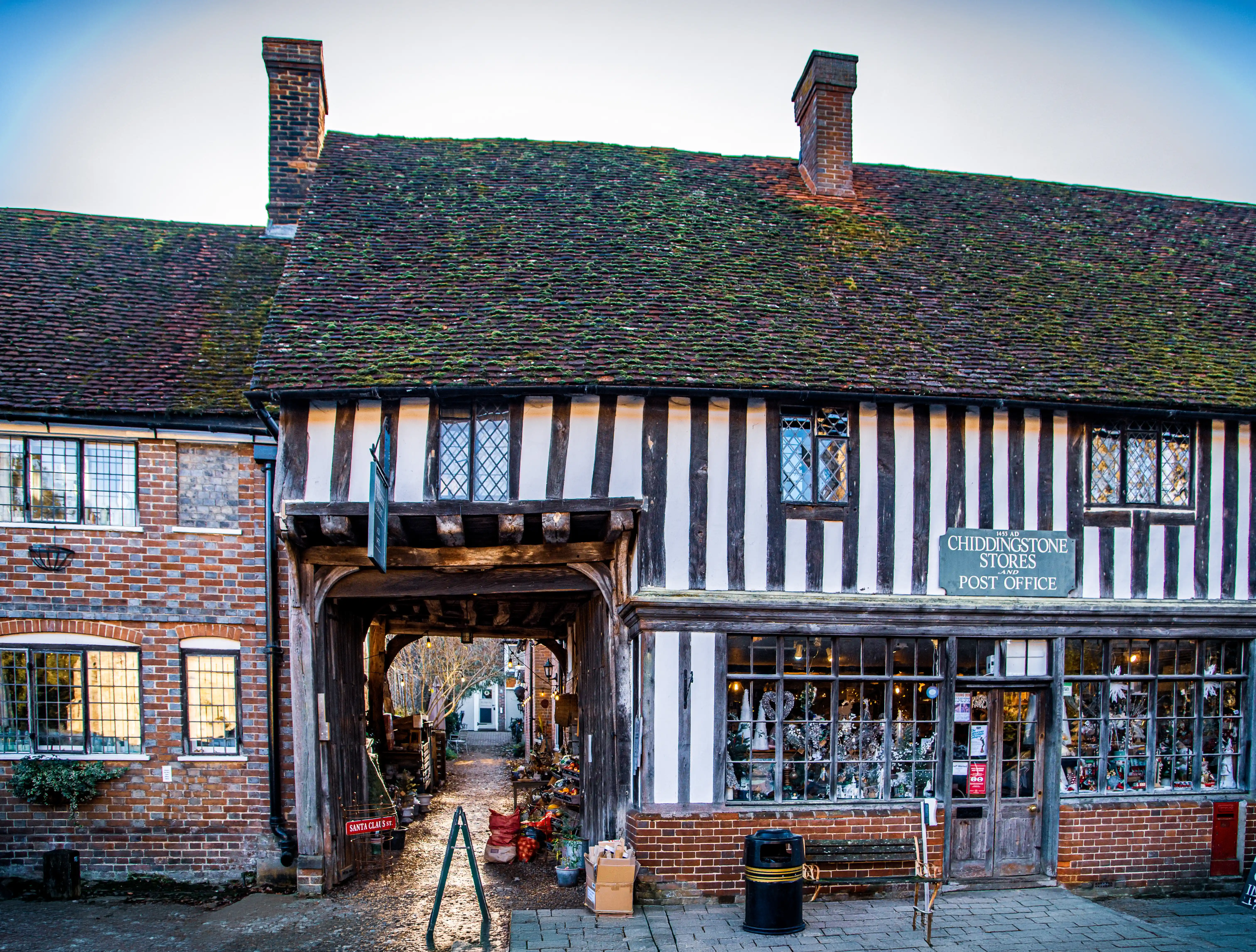Exterior view of the Chiddingstone Stores and Post Office, a historic building with black and white timber framing, a tiled roof, and leaded glass windows. An archway leads to a village street, and a sign indicates 