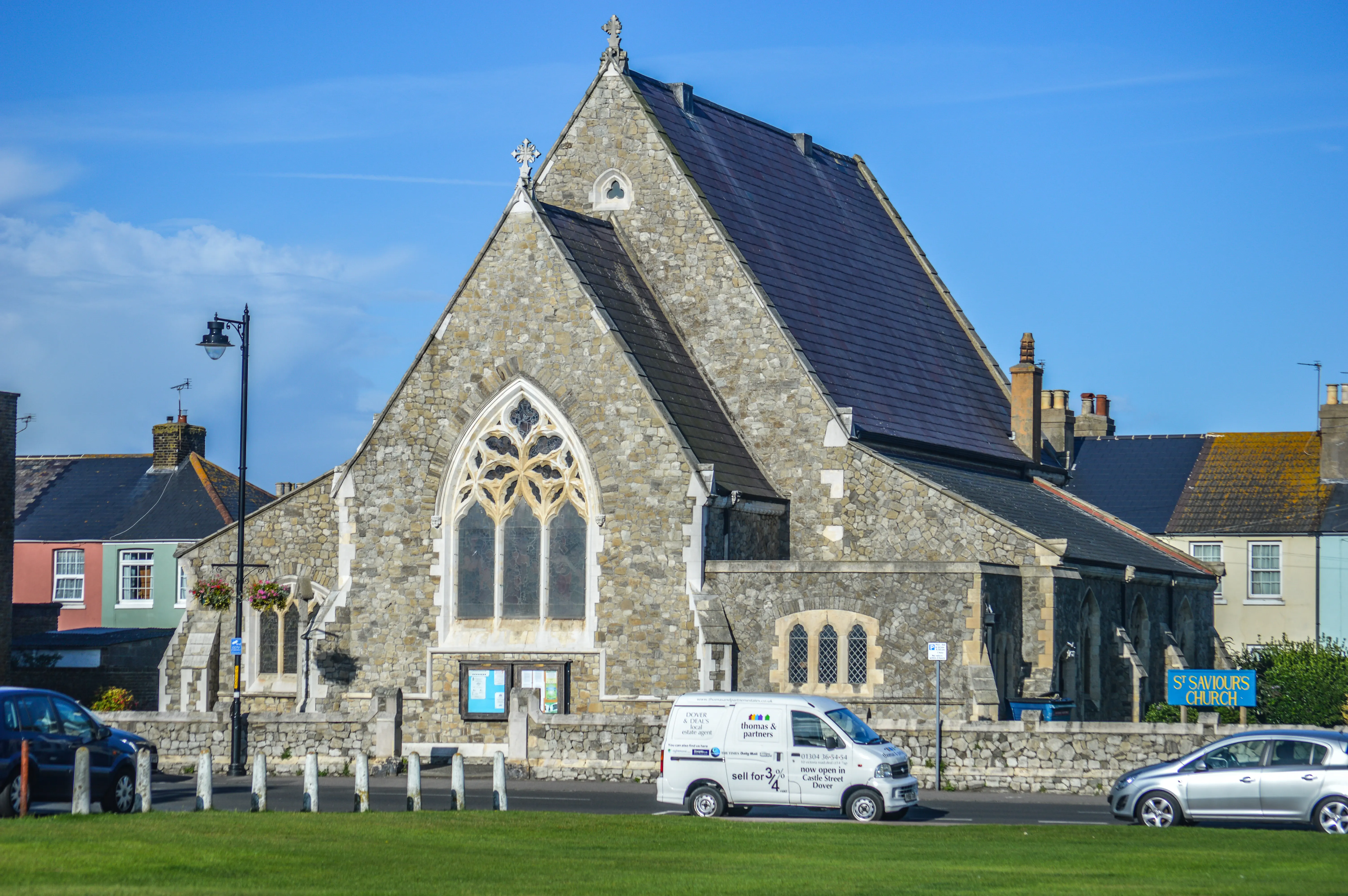 White estate agent van parked outside St Saviour's Church, with a silver car driving past on the road and a dark blue car parked to the left. The church is made of stone with a large arched window.