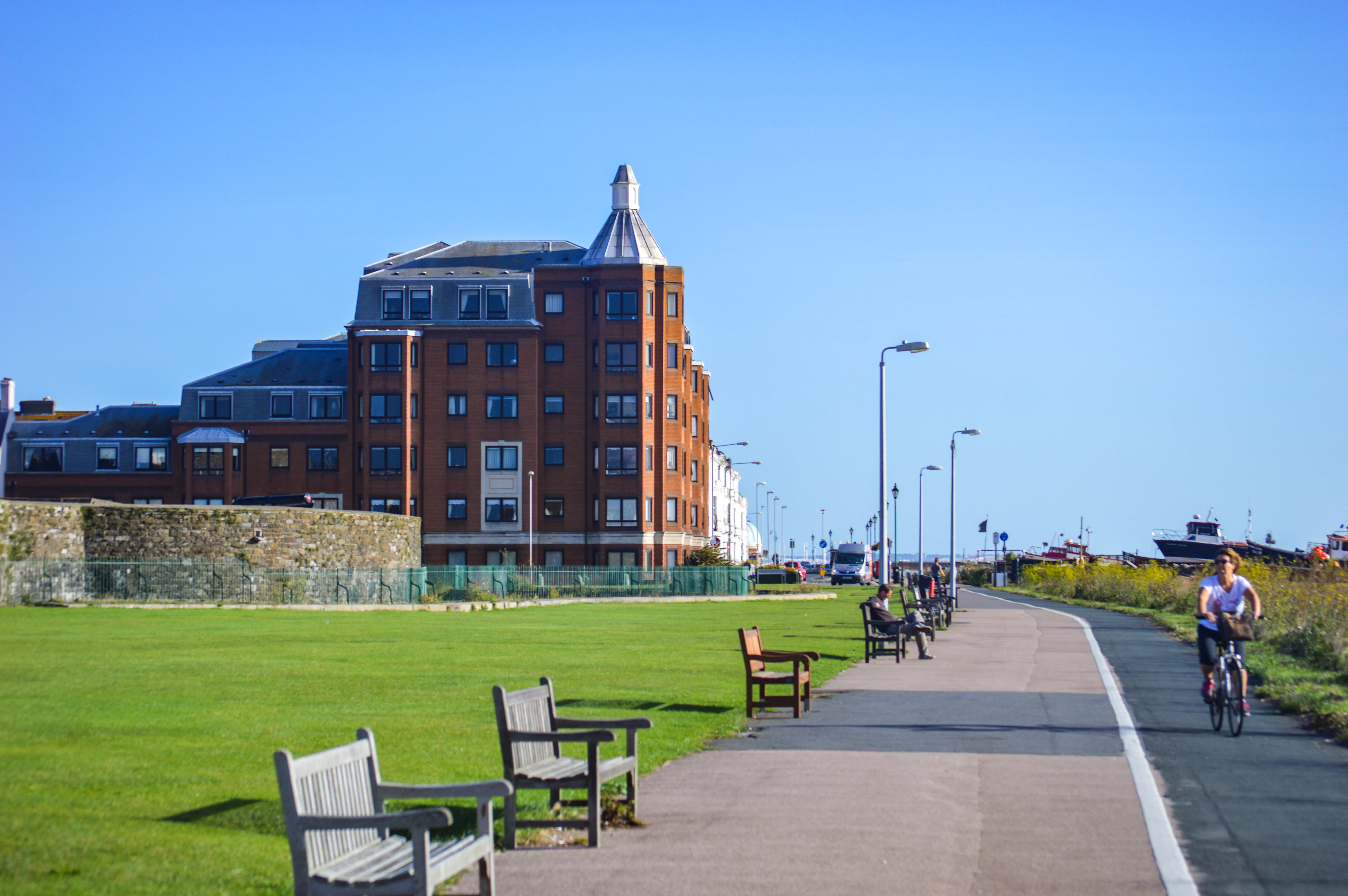 A wide shot of a coastal promenade on a clear day. On the left is a large, multi-story brick building with many windows. In front of it is a well-maintained lawn with several empty wooden benches. A paved path runs along the right side of the lawn, where a woman is cycling and a few people are sitting on benches. In the background, beyond the path, are several boats docked and a line of streetlights.