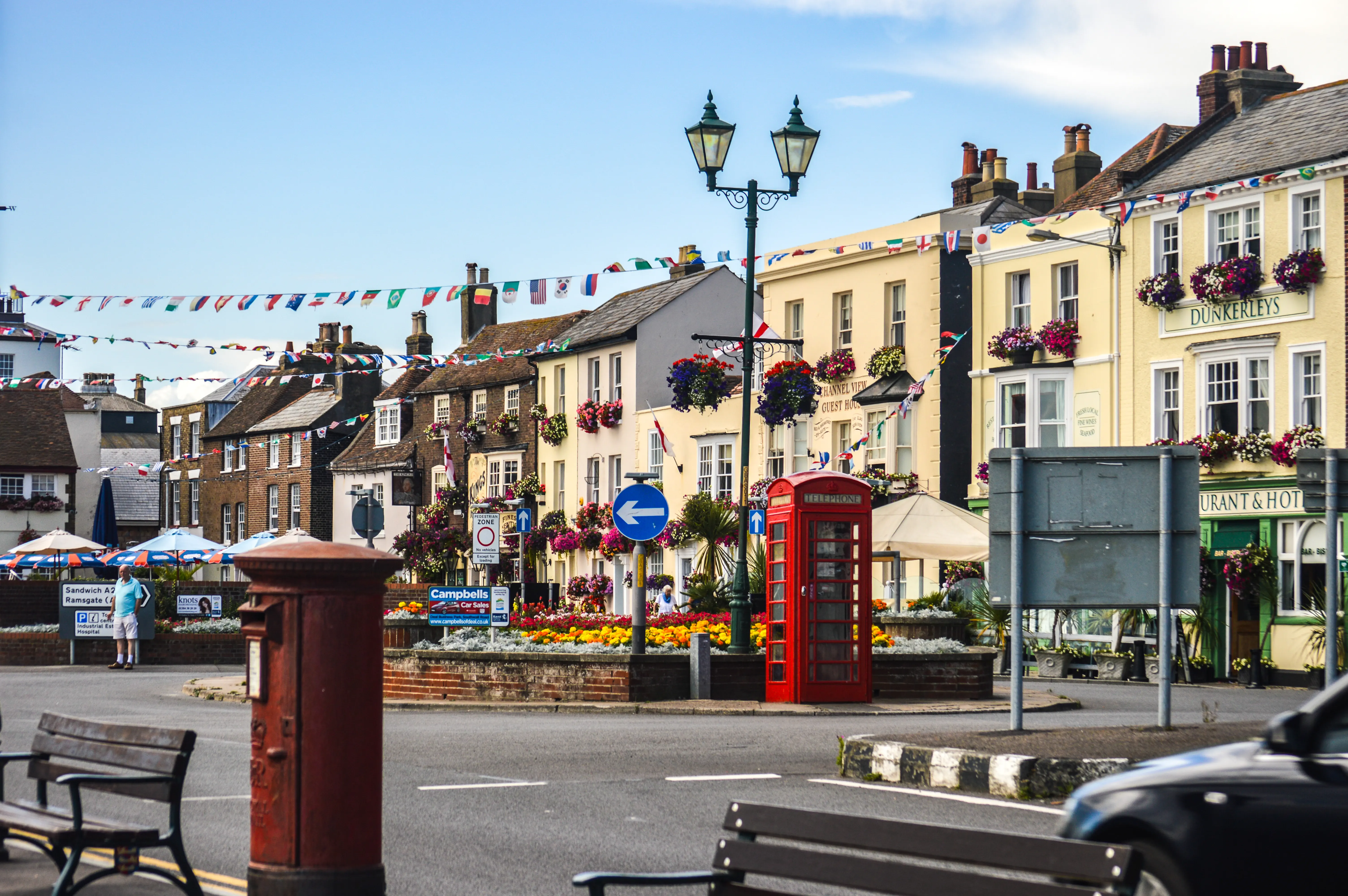 A red telephone box is situated on a street lined with buildings decorated with colourful flags and hanging flower baskets. A person is visible walking in the background, and a black car is partially seen in the foreground.