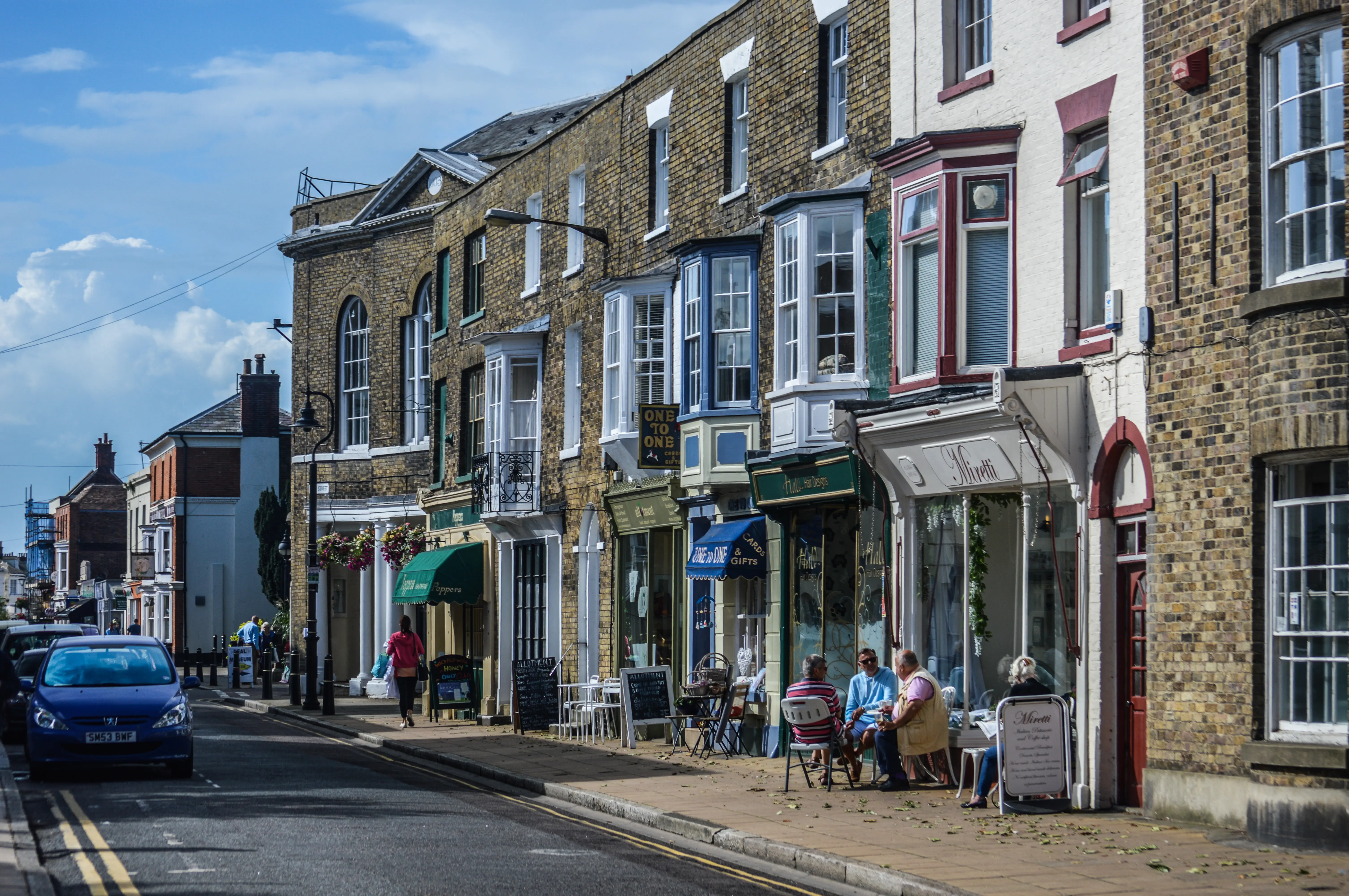 A street view of a row of traditional brick shops with bay windows, people sitting outside a cafe, and a blue car parked on the road under a blue sky.