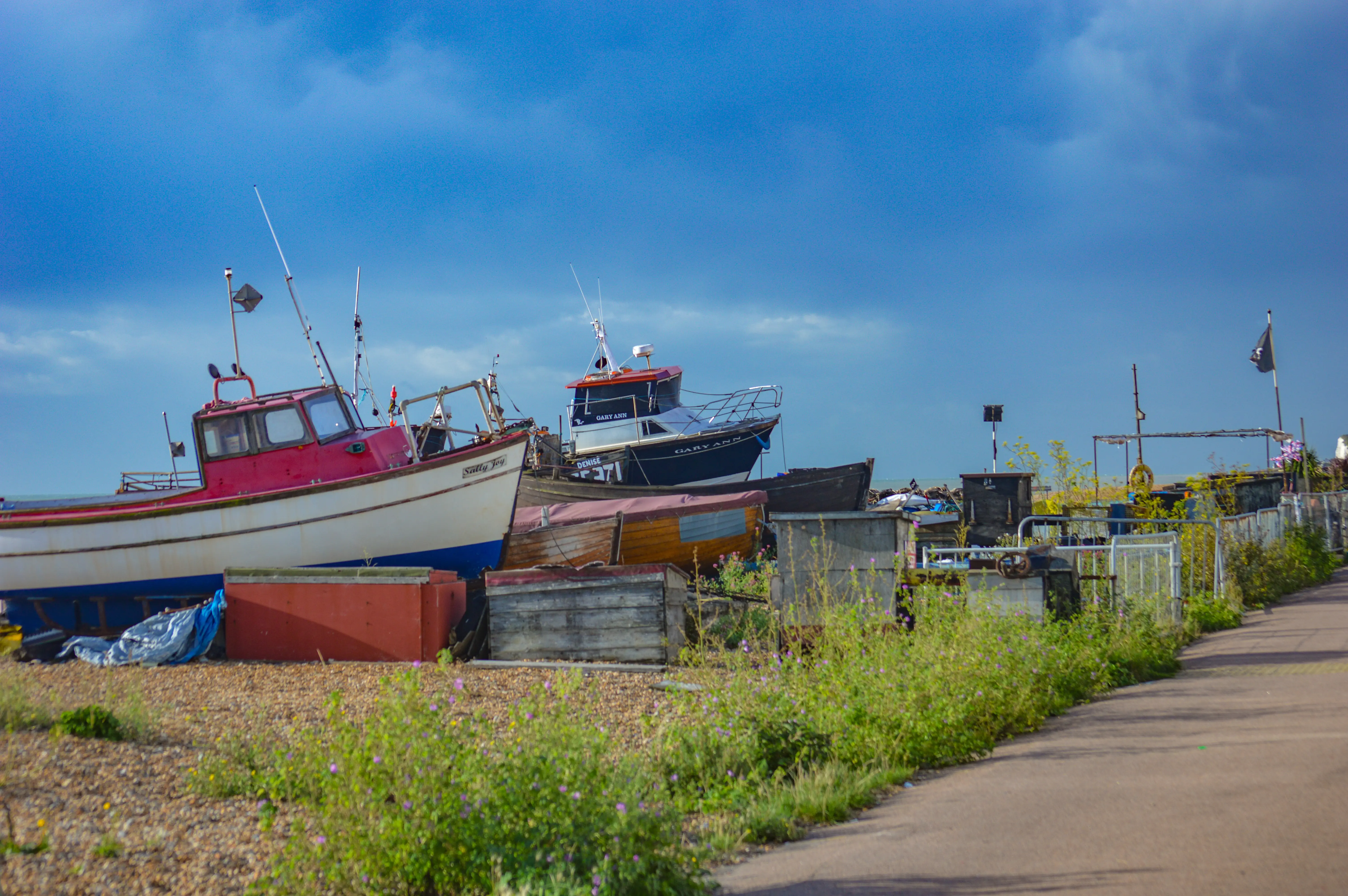 Fishing boats of various sizes are pulled up onto a shingle beach. The closest boat is white with a red cabin and the name Salty Joy visible. Behind it are other boats, including one with Gary Ann written on its hull. A paved path runs along the right side of the image, bordered by overgrown greenery. A black pirate flag flutters in the distance against a greyish-blue sky.