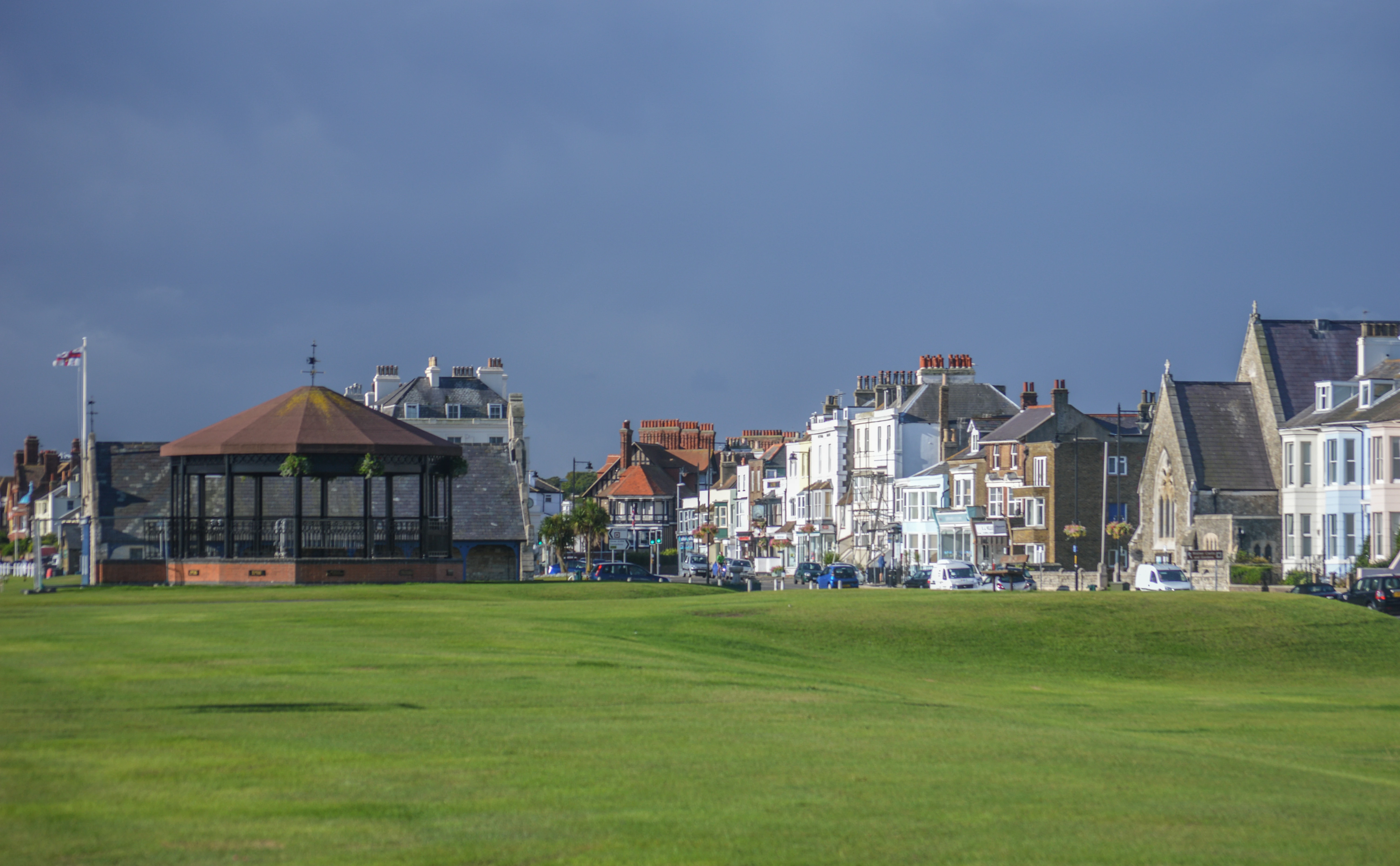 A wide shot of a British seaside town with a green lawn in the foreground and a bandstand. A line of residential and commercial buildings stretches along the coast, with a dramatic, dark sky overhead.