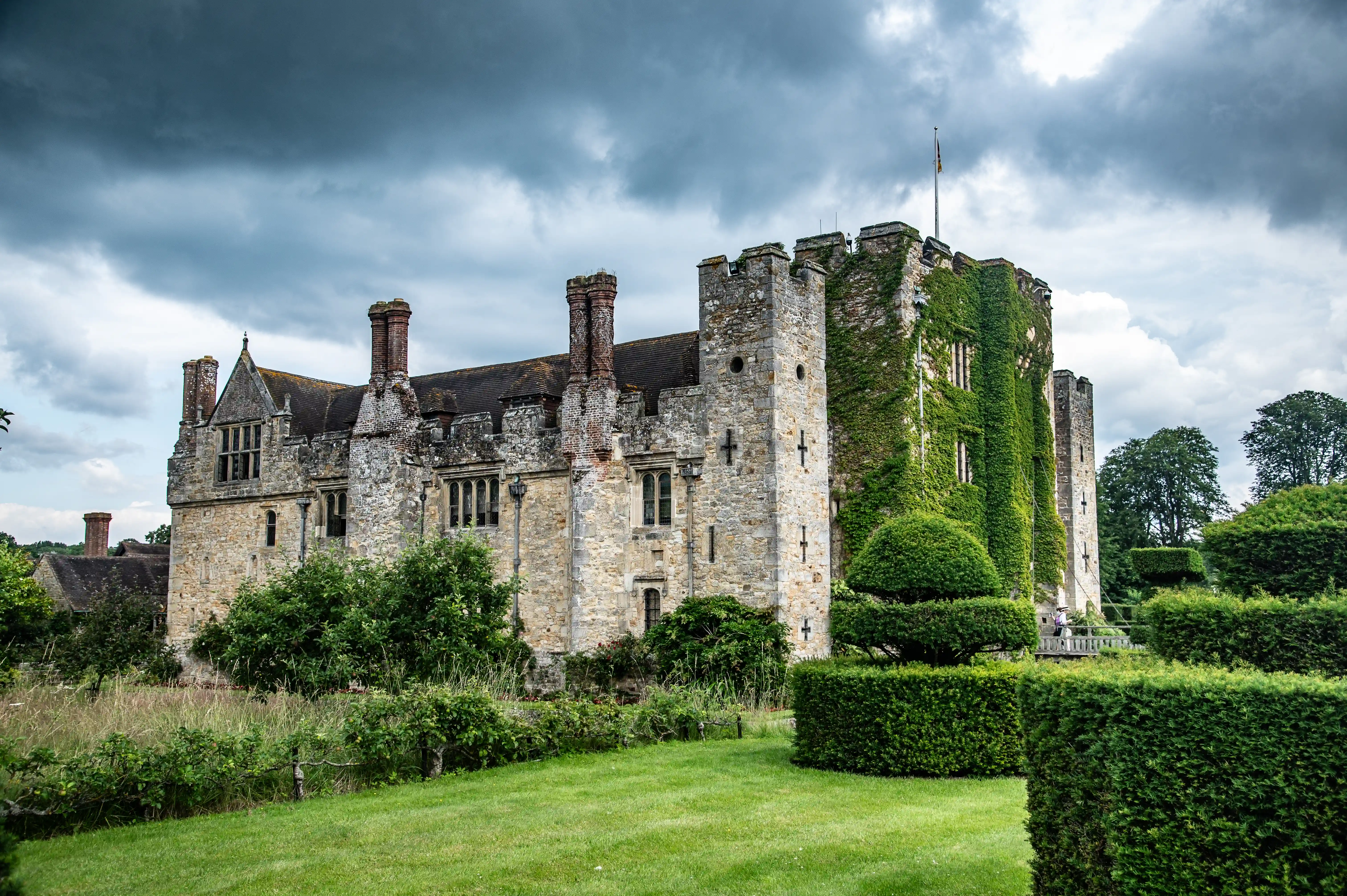 A stone castle with ivy climbing its walls, set against a stormy sky, with formal gardens in the foreground.