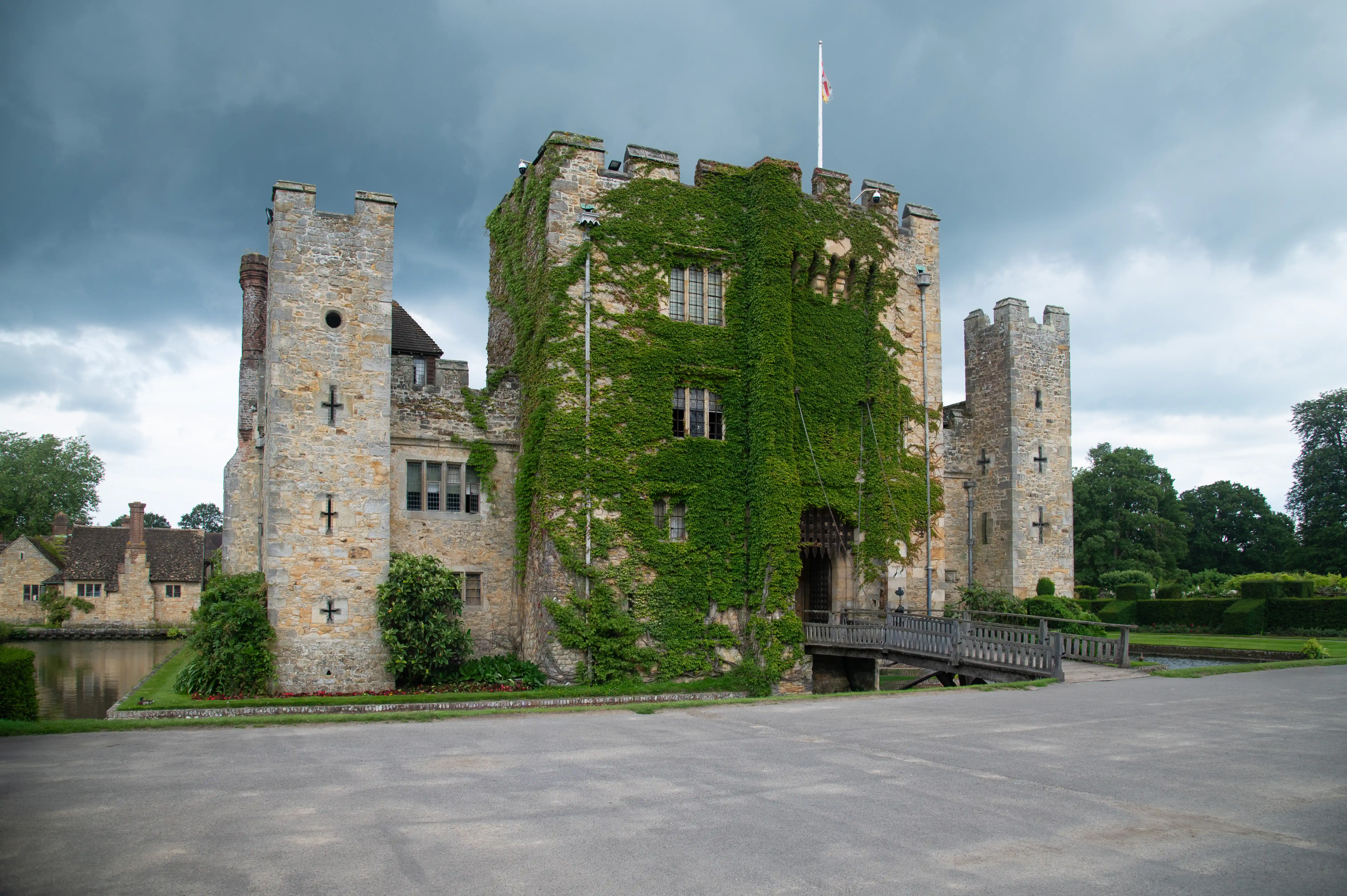 A historic stone castle with a drawbridge and ivy covering its walls, set against a dark, cloudy sky, with a pond and gardens in the foreground.