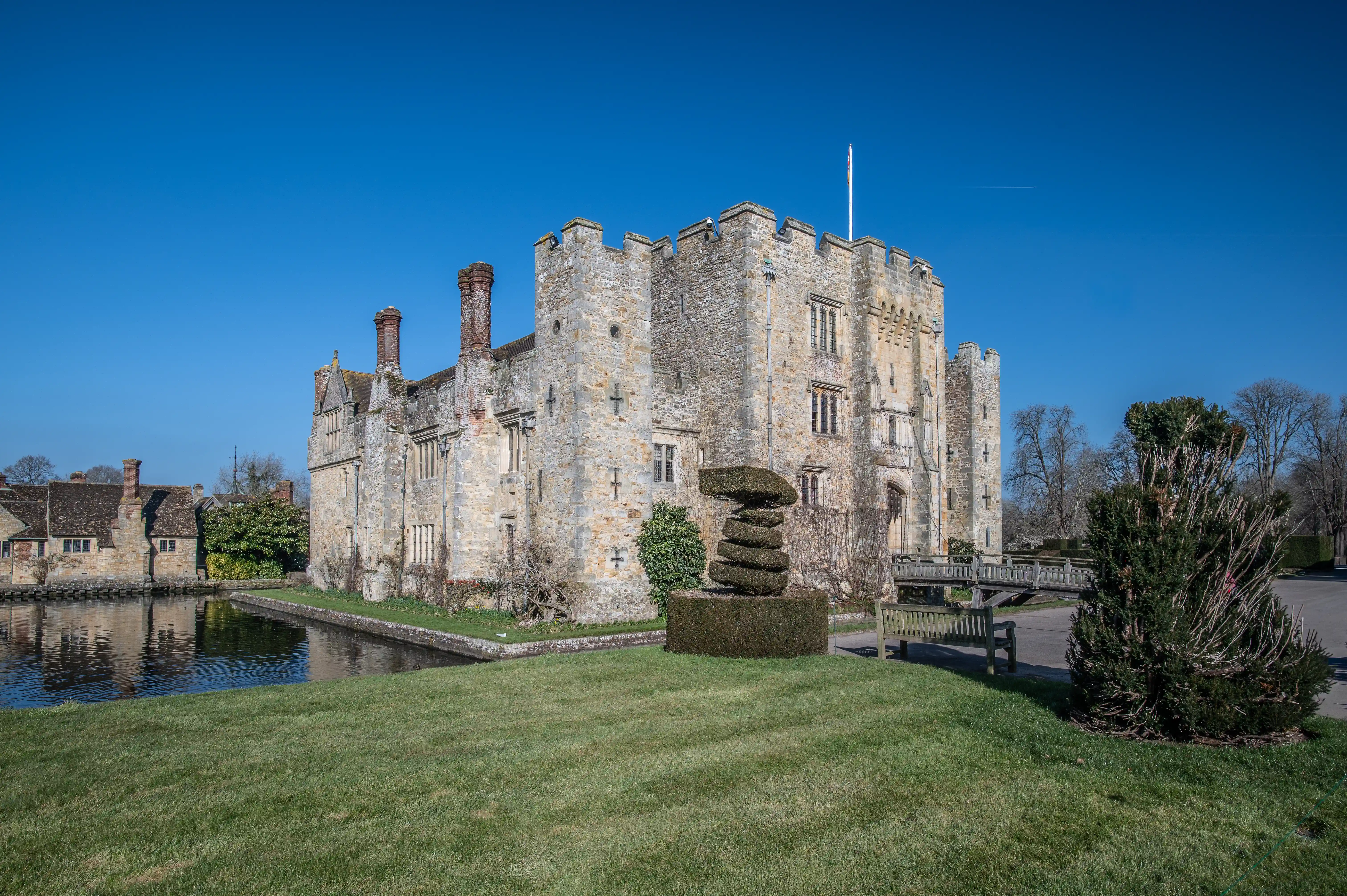 A wide view of a historical stone castle with a moat in front, a formal garden featuring a unique stacked hedge sculpture, and a wooden bridge, under a clear blue sky.