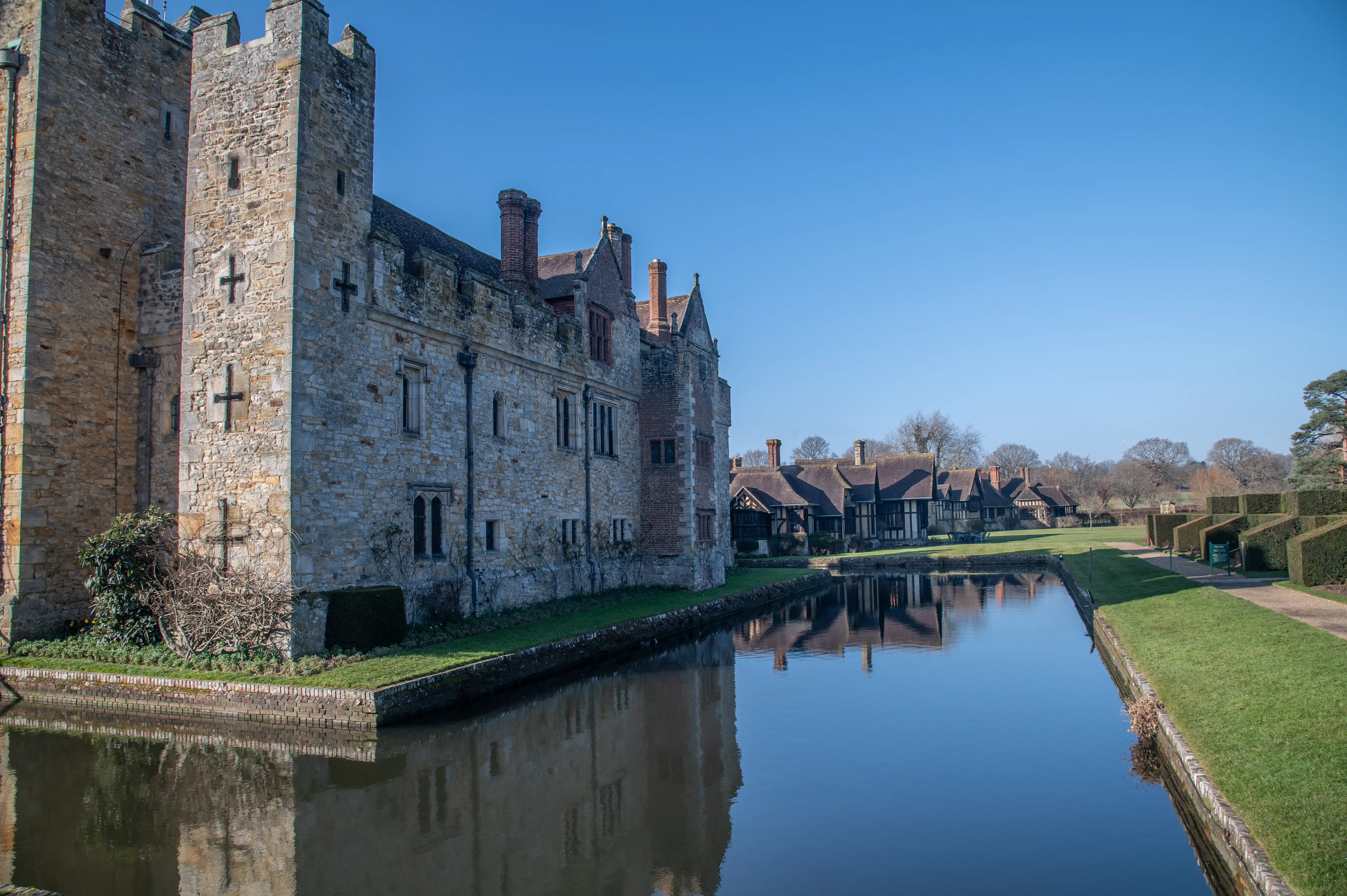 A wide-angle view of a historic stone castle with a moat. The castle's reflection is visible in the water. In the background, a line of traditional Tudor-style houses with dark timber and white walls is set against a clear blue sky.