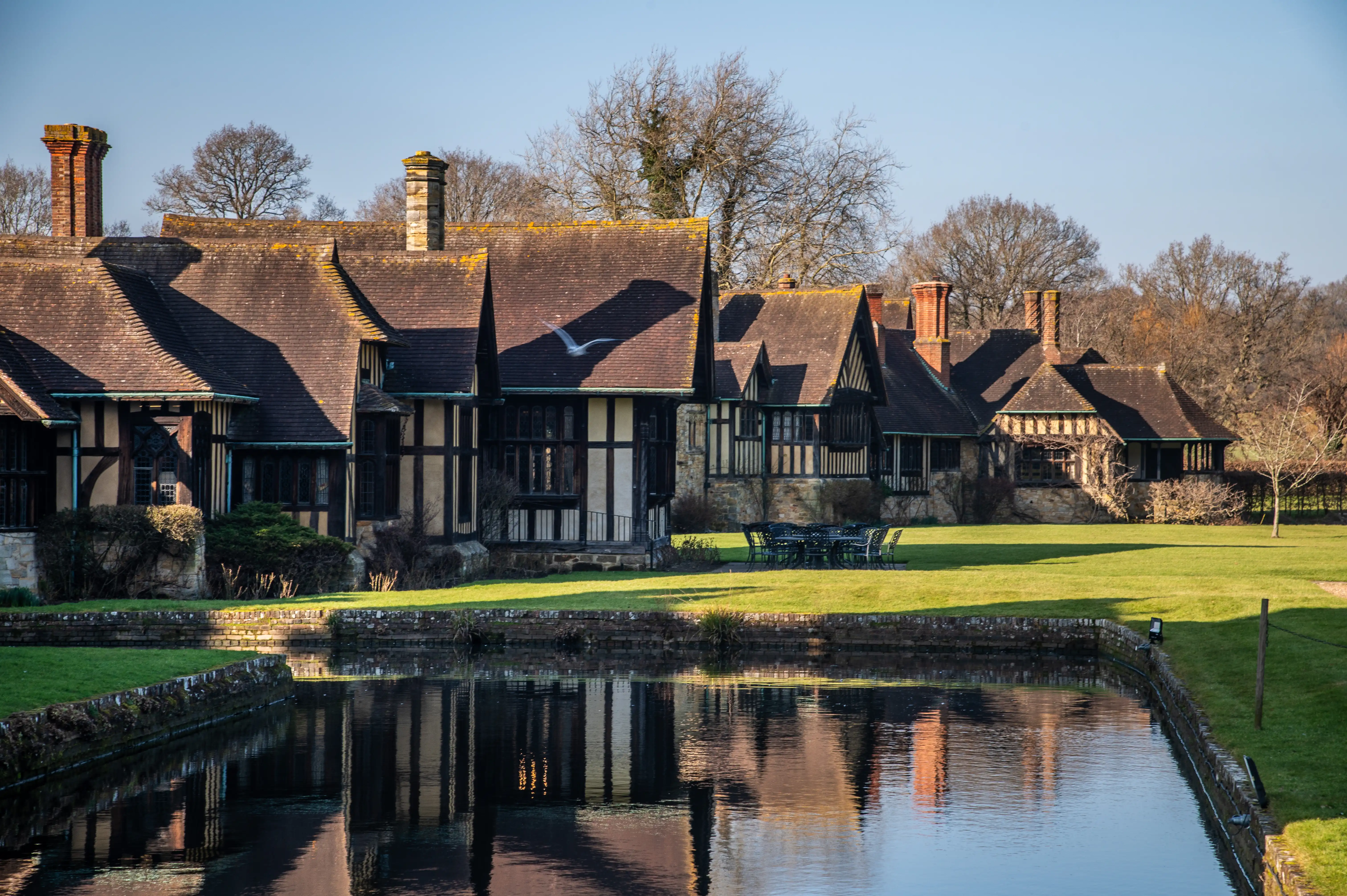 A long, multi-gabled Tudor manor house with dark timber framing and cream infill, seen across a still pond reflecting the building and the bright blue sky. There is a garden table and chairs on the manicured lawn.