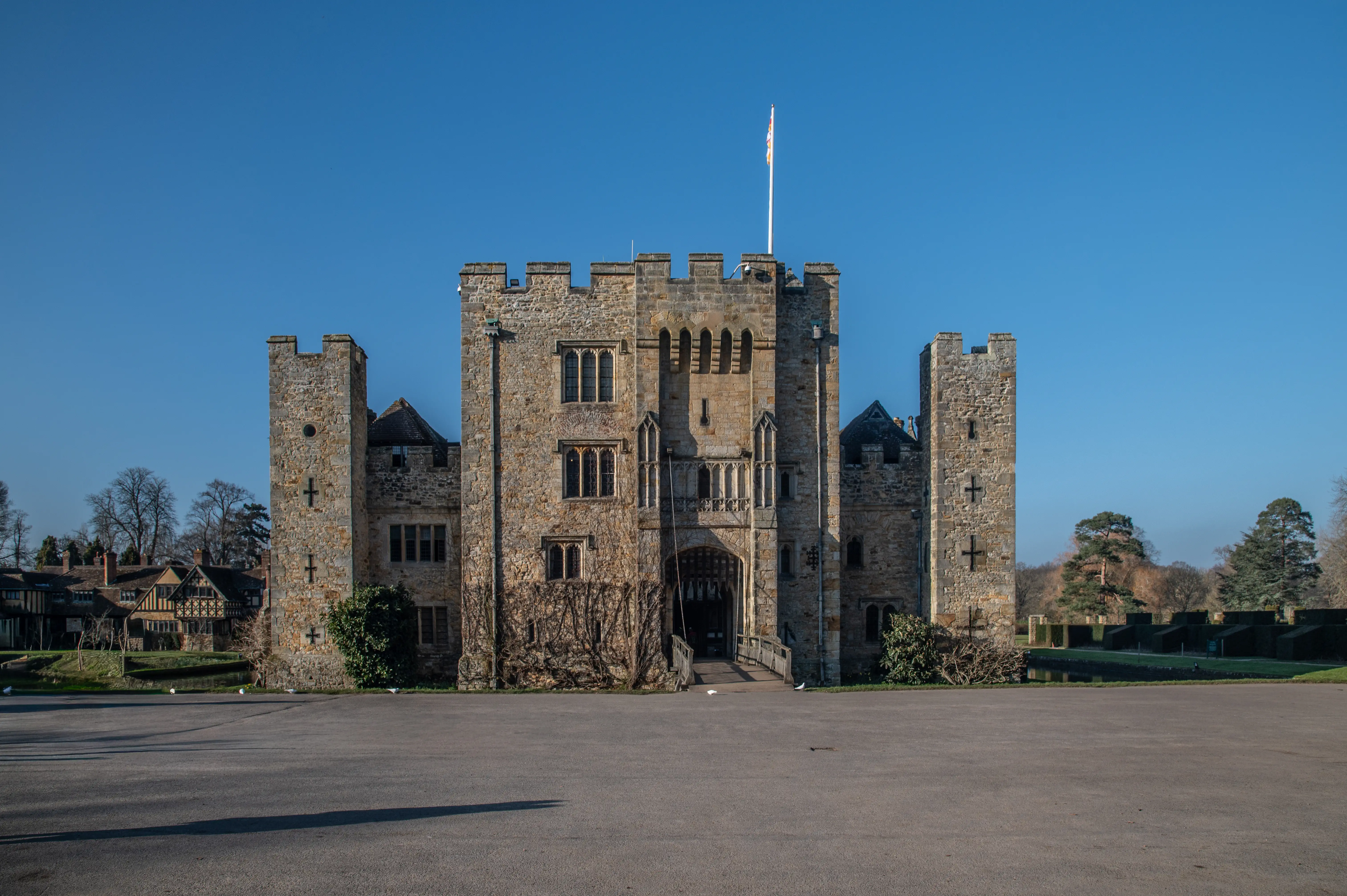 A photograph of a large, imposing stone castle with multiple towers and a central entrance reached by a wooden bridge. The castle is set against a bright blue, cloudless sky, and the foreground is a flat, dark paved area.