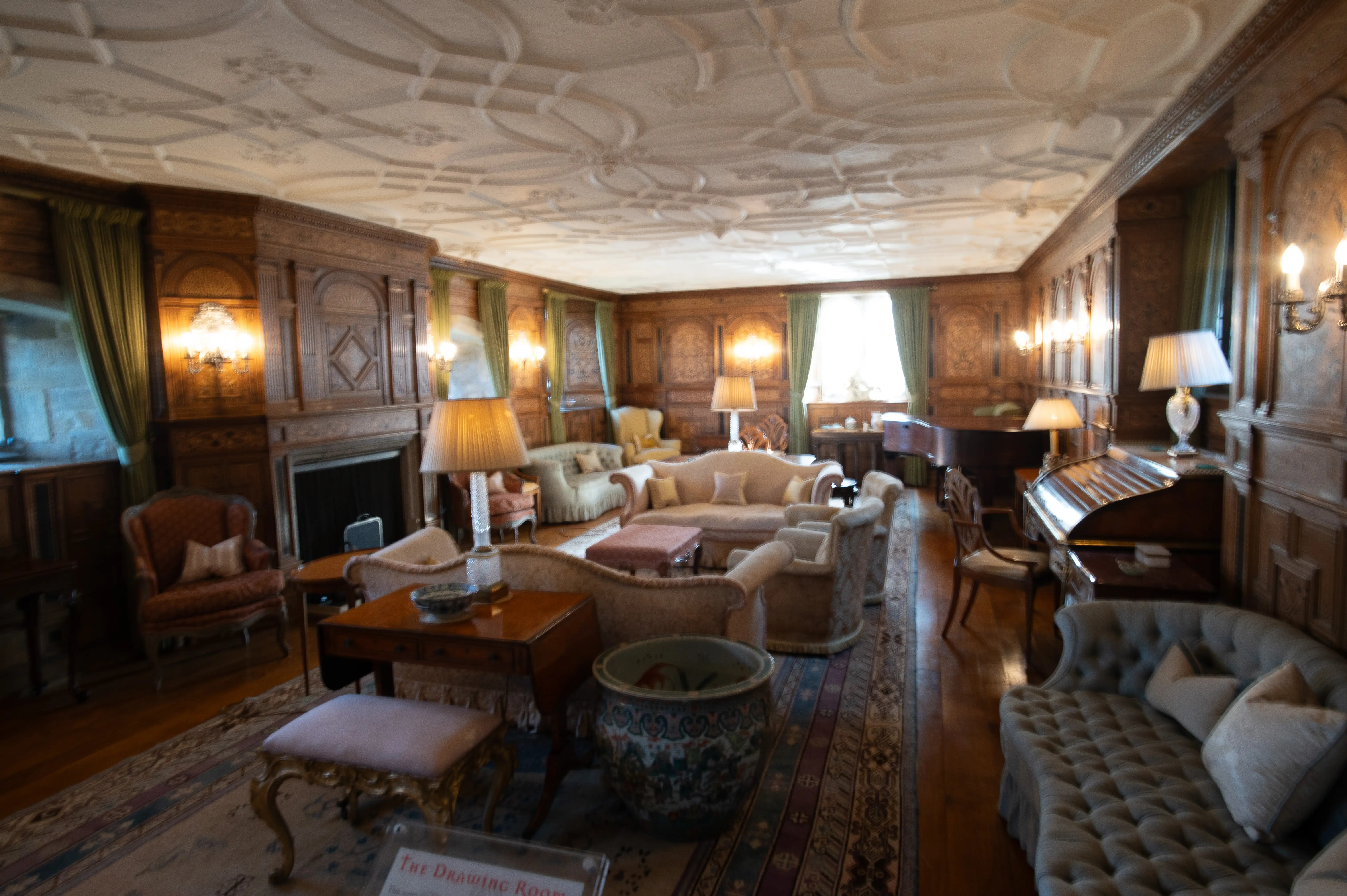 A wide-angle view of a luxurious drawing room featuring dark wood panelling, a detailed plaster ceiling, several sofas and armchairs, a fireplace, a grand piano, and lamps.