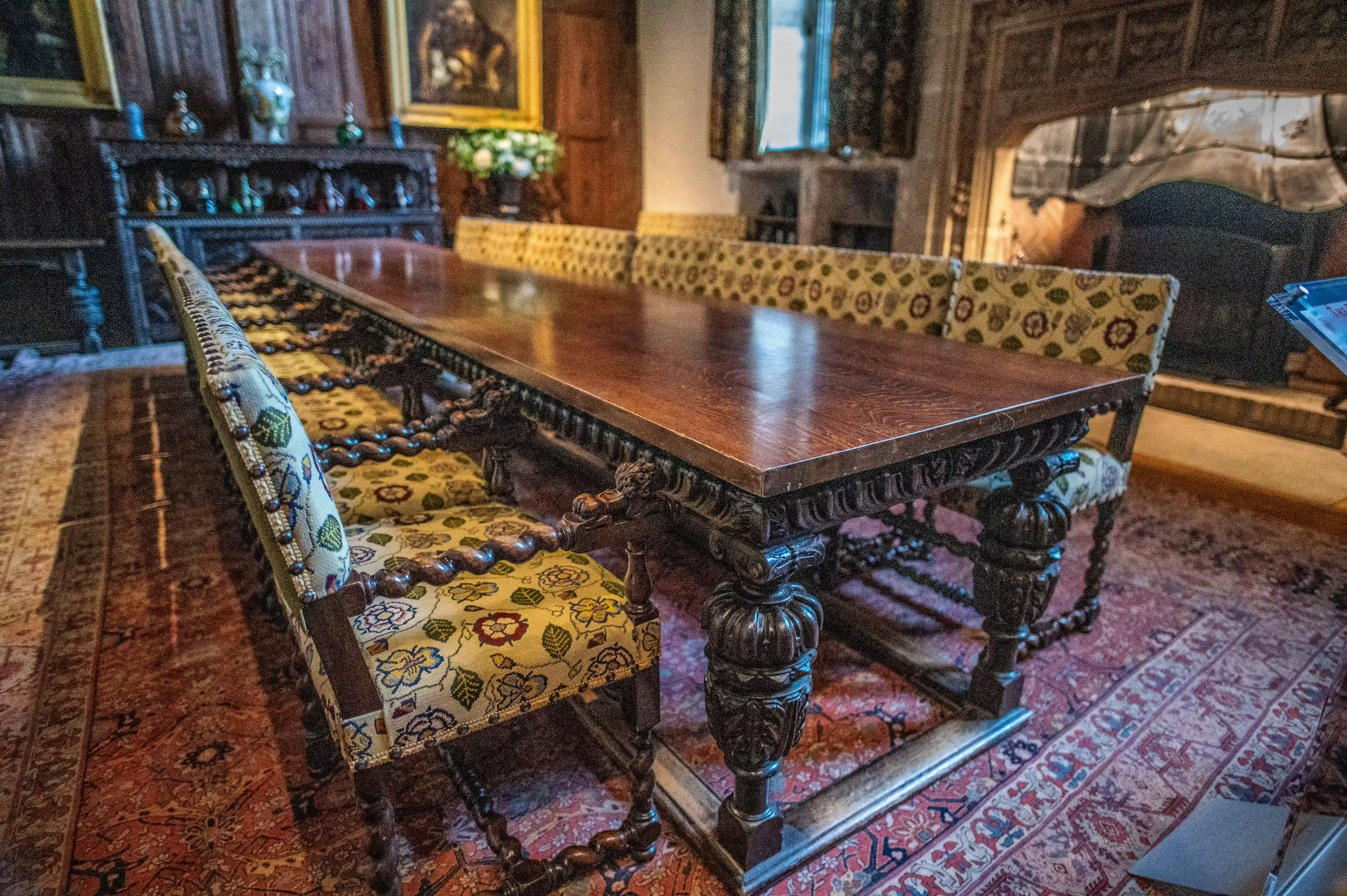 A long, ornately carved dark wood dining table with matching chairs upholstered in floral fabric, set in a grand dining hall with a fireplace and antique decor.