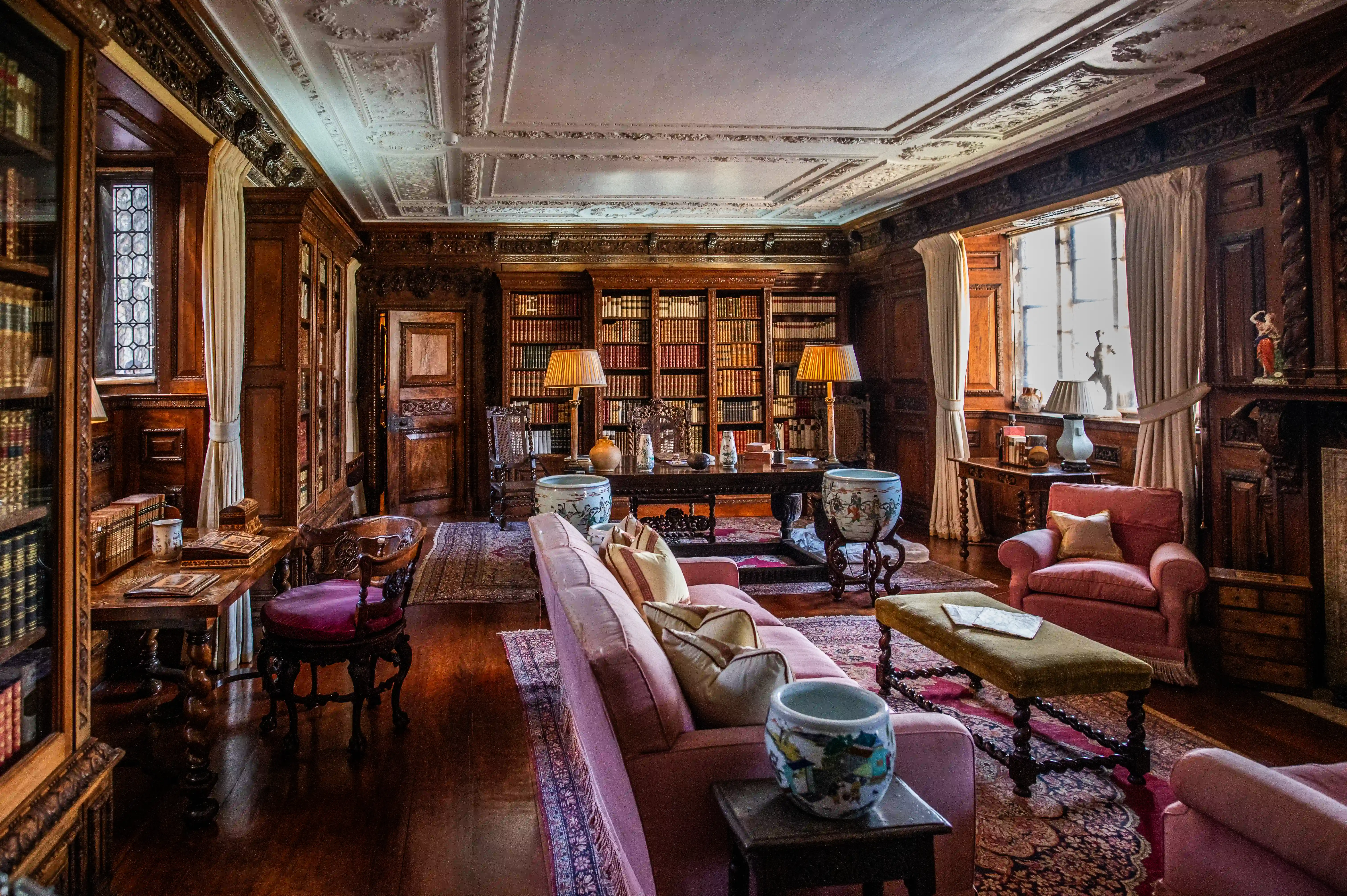 This image captures a beautifully furnished drawing room, likely within a historic estate or stately home. The room is adorned with rich wood panelling, an ornate coffered ceiling, and plush seating arrangements. A grand piano, a fireplace, and period furniture contribute to the room's opulent atmosphere.