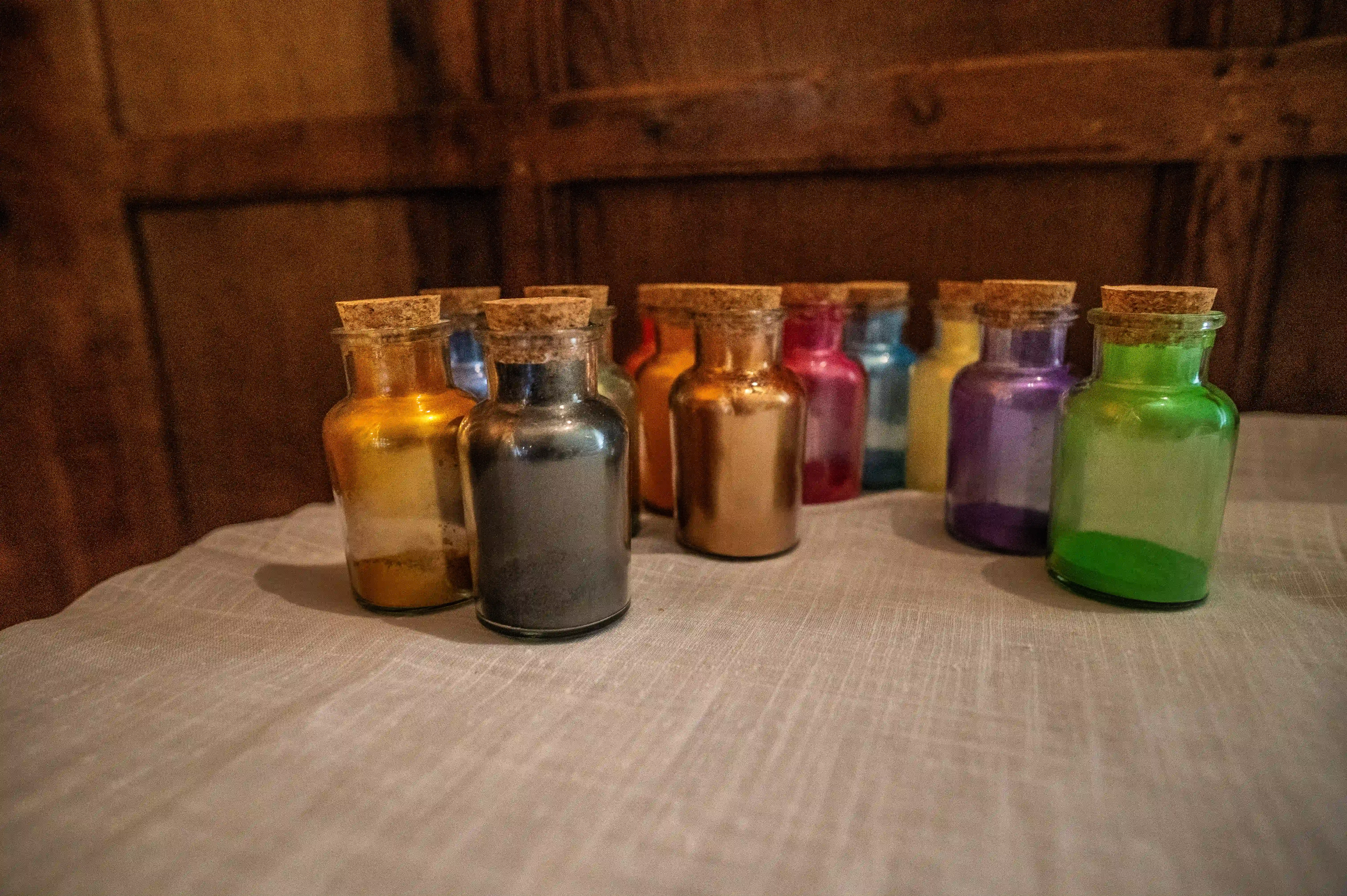 Several small glass bottles filled with coloured powders like gold, grey, bronze, pink, blue, purple, and green, arranged on a textured surface against a wooden background.