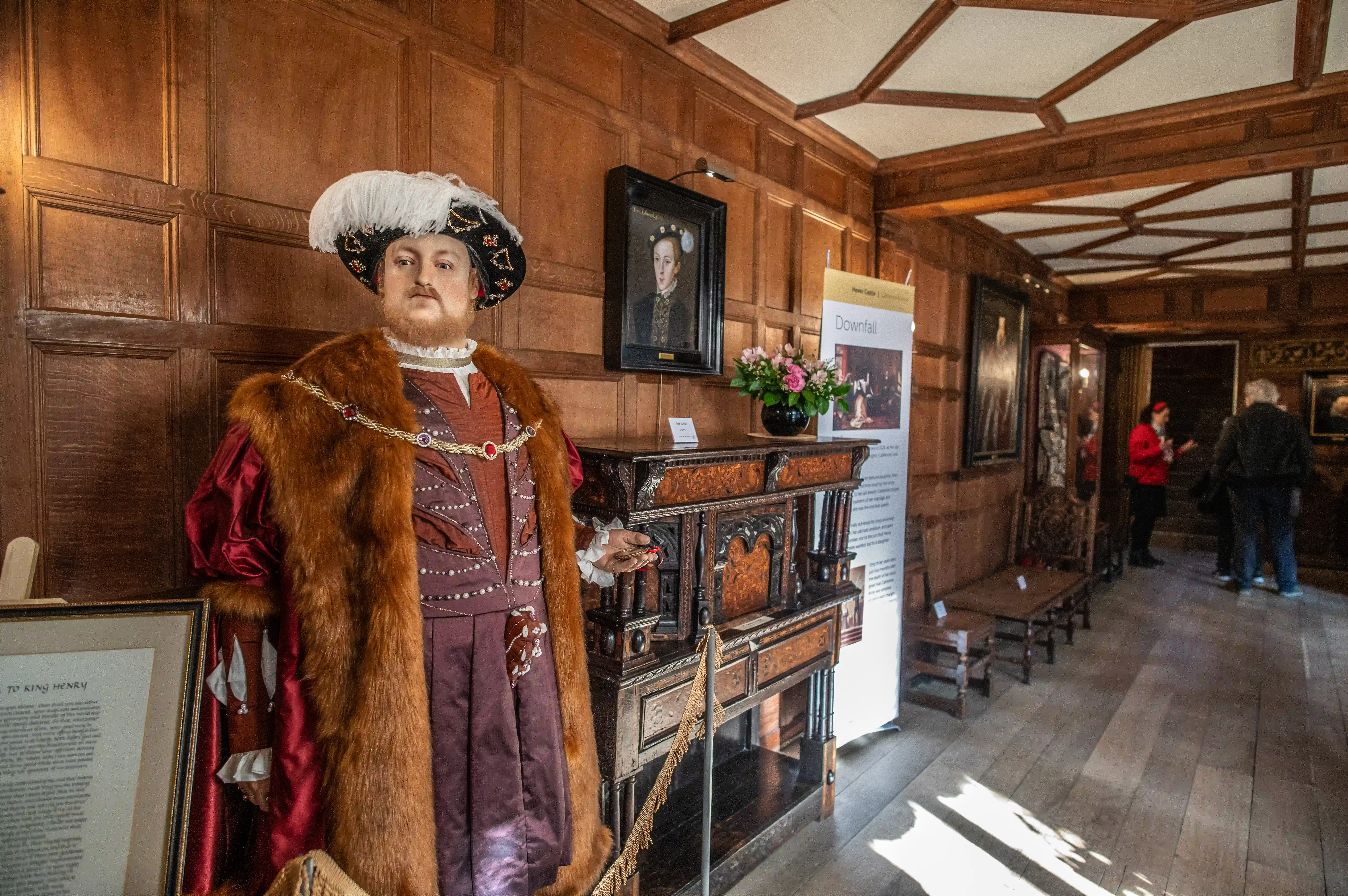 Mannequin of King Henry VIII in historical costume in a museum exhibition room, with wood panelling, antique furniture, and visitors in the background.