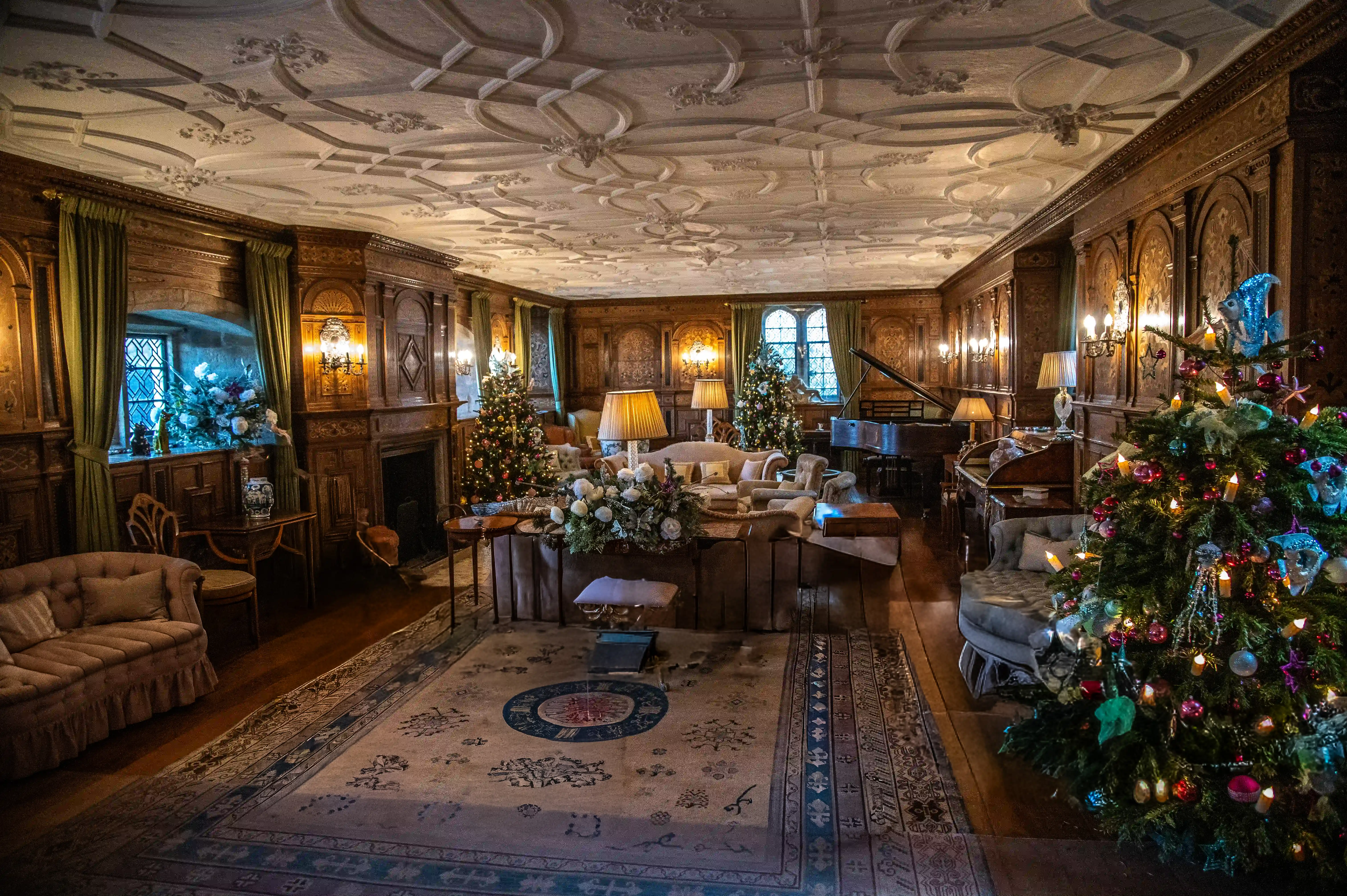 A lavishly decorated drawing-room in a historic home, featuring Christmas trees, ornate wood panelling, a grand piano, and plush furniture.