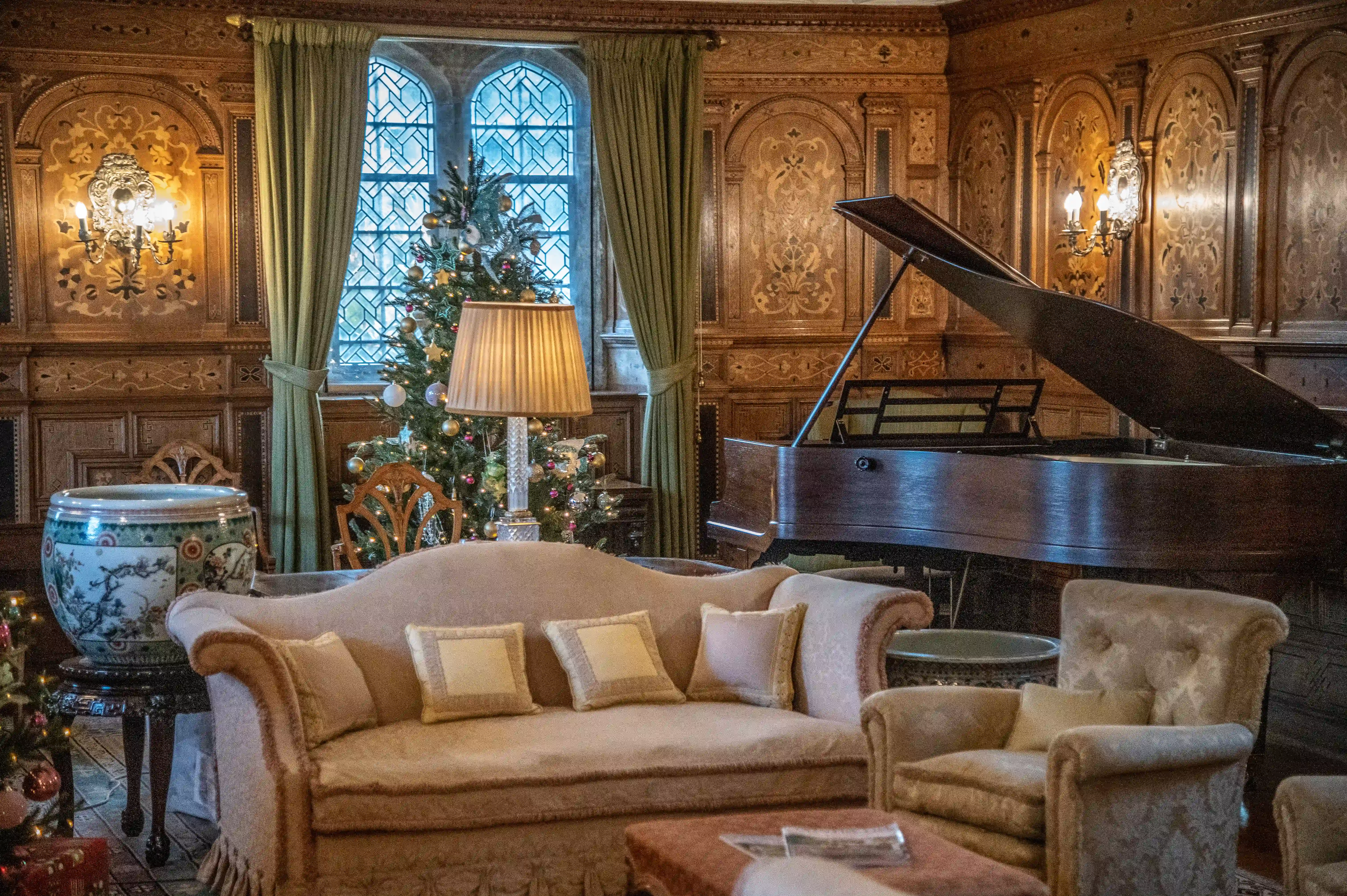 A dimly lit, wood-panelled drawing-room decorated for Christmas, with a grand piano, a sofa, armchairs, and a Christmas tree next to a large gothic-style window.