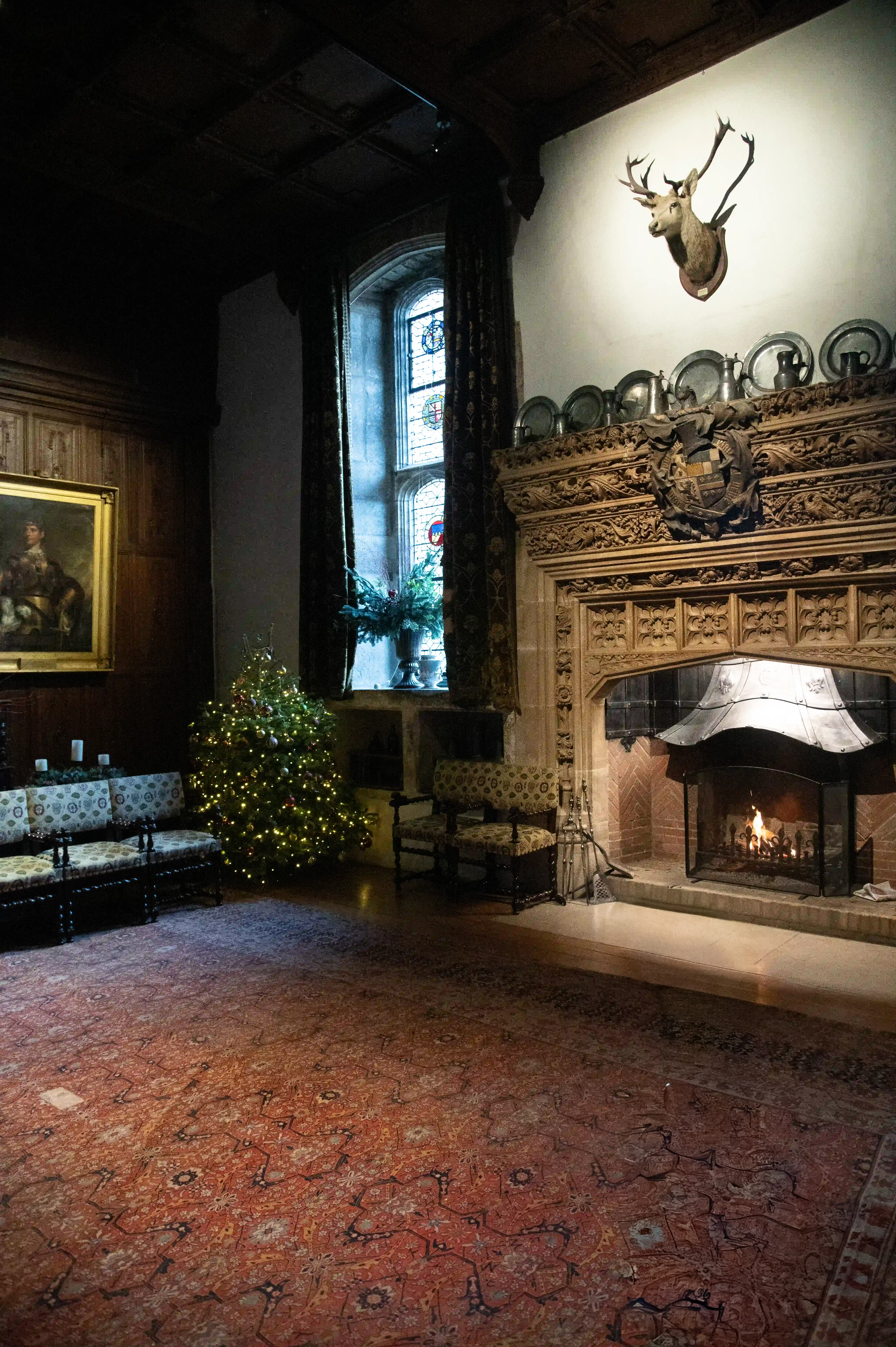 A dimly lit, grand drawing room is set for Christmas. In the foreground, a large, ornate Persian rug covers the floor. To the left, a Christmas tree decorated with lights and baubles stands near two upholstered benches. Opposite the tree, a large, carved stone fireplace with a lit fire is visible, topped by a mounted stag's head. A large stained glass window is to the left of the fireplace.