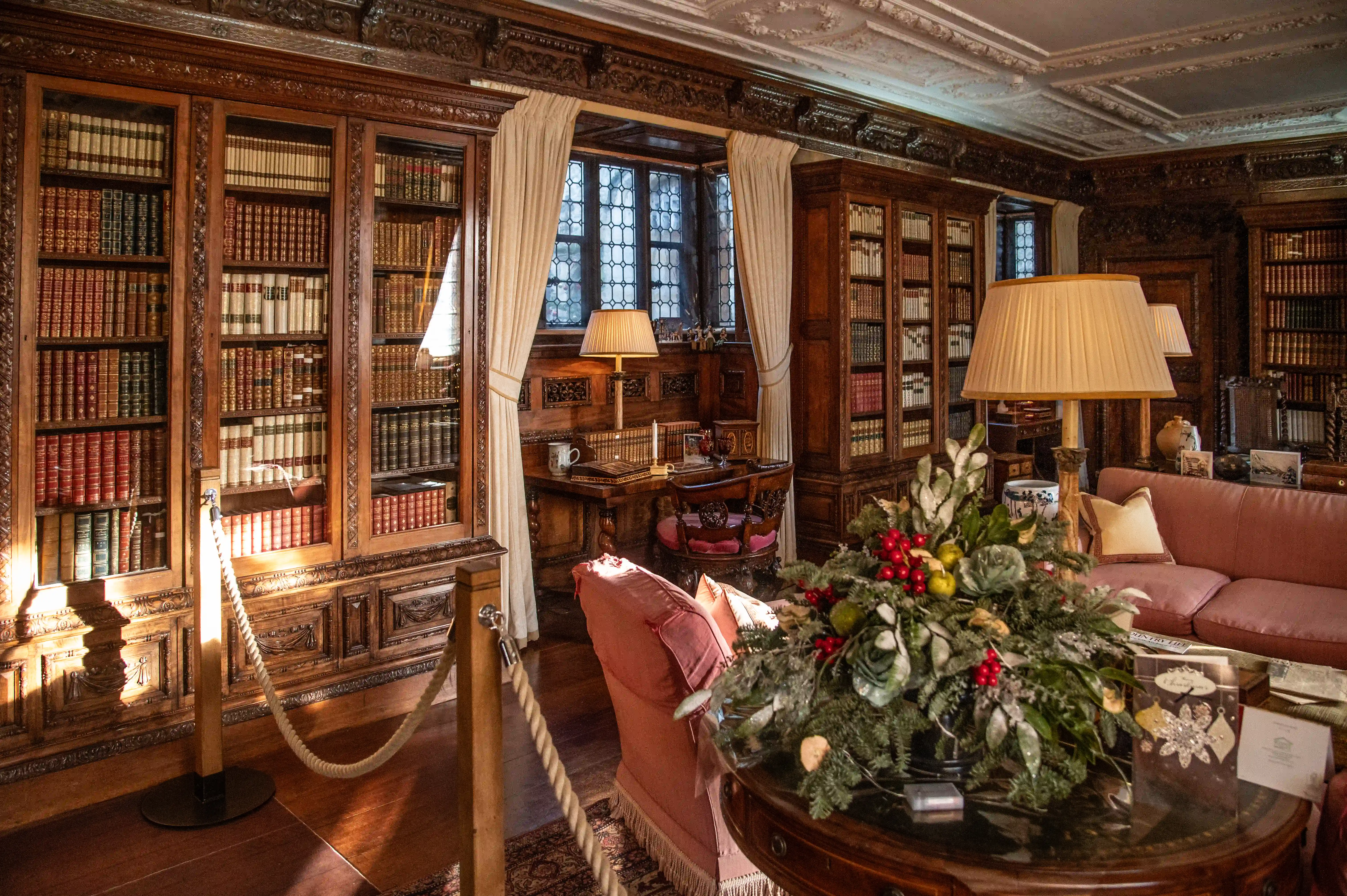 A wide shot of a luxurious library with dark wood bookcases filled with books, a pink sofa, a desk by a bay window, and a festive floral arrangement in the foreground. Sunlight streams in from the left.