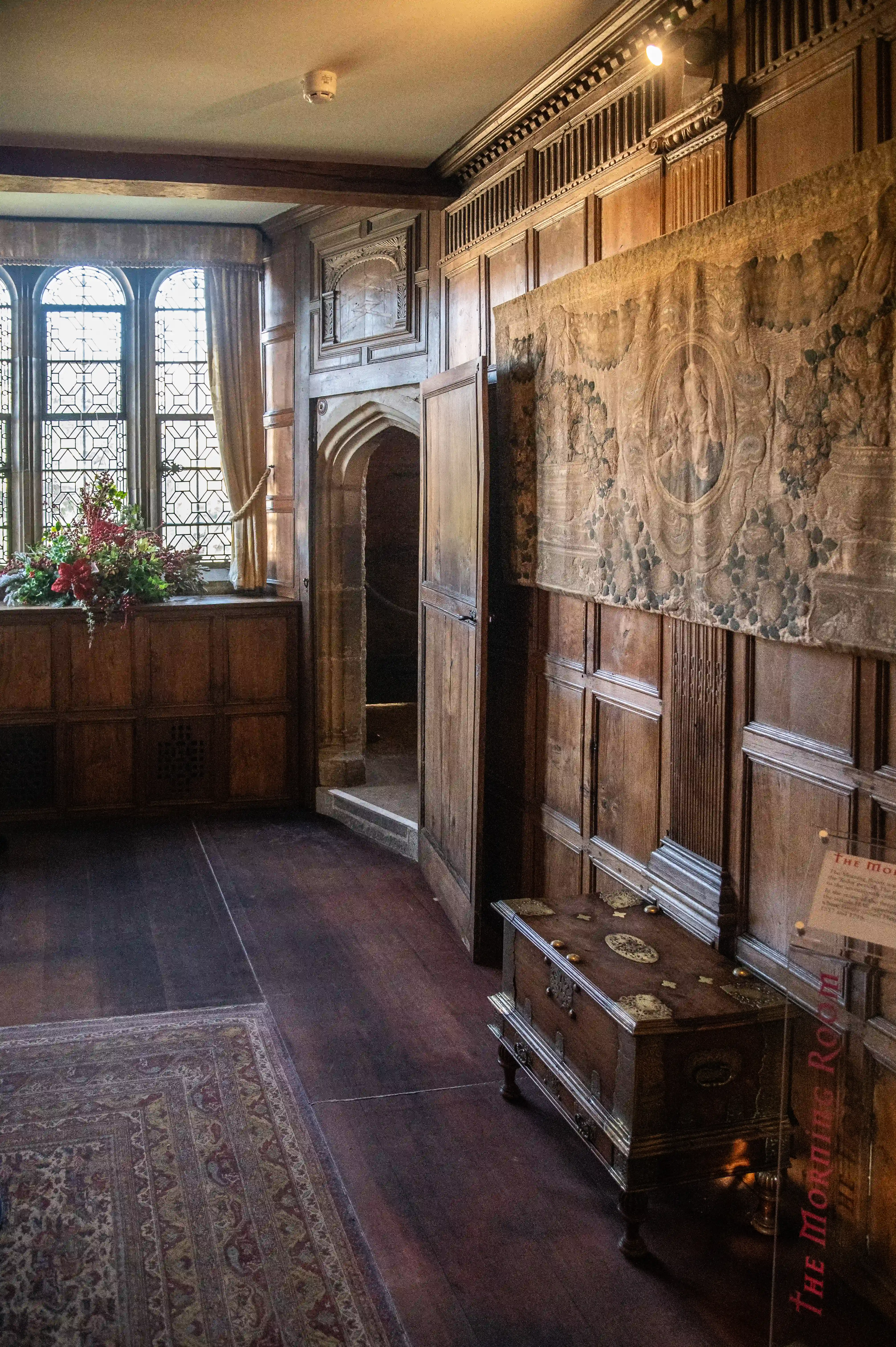 Interior view of a historic room with wood panelling, a tapestry, a decorative chest, a leaded glass window with floral arrangement, and an arched doorway.