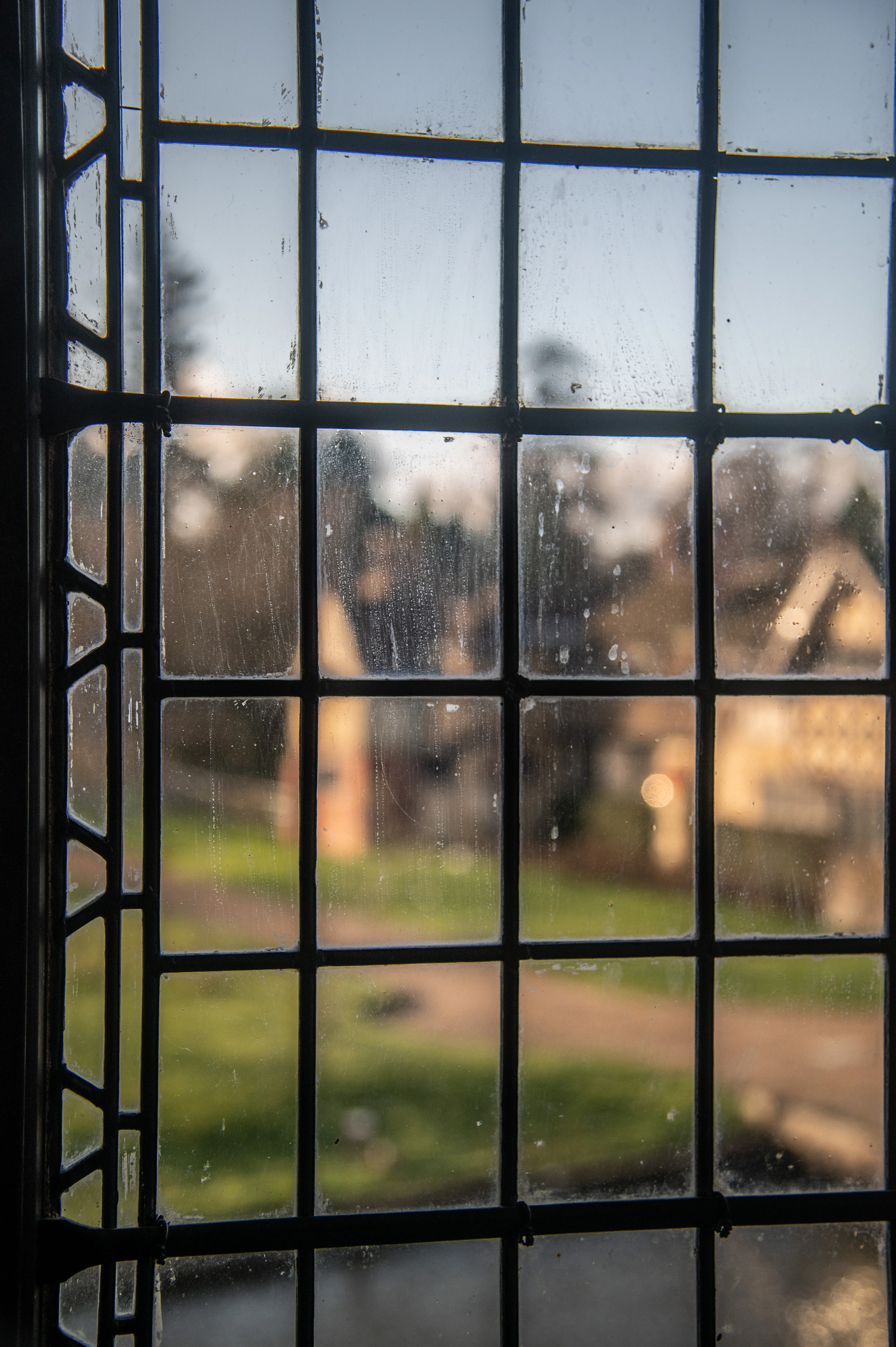 View through a leaded glass window with water droplets and smudges, showing a blurred landscape with houses and greenery.