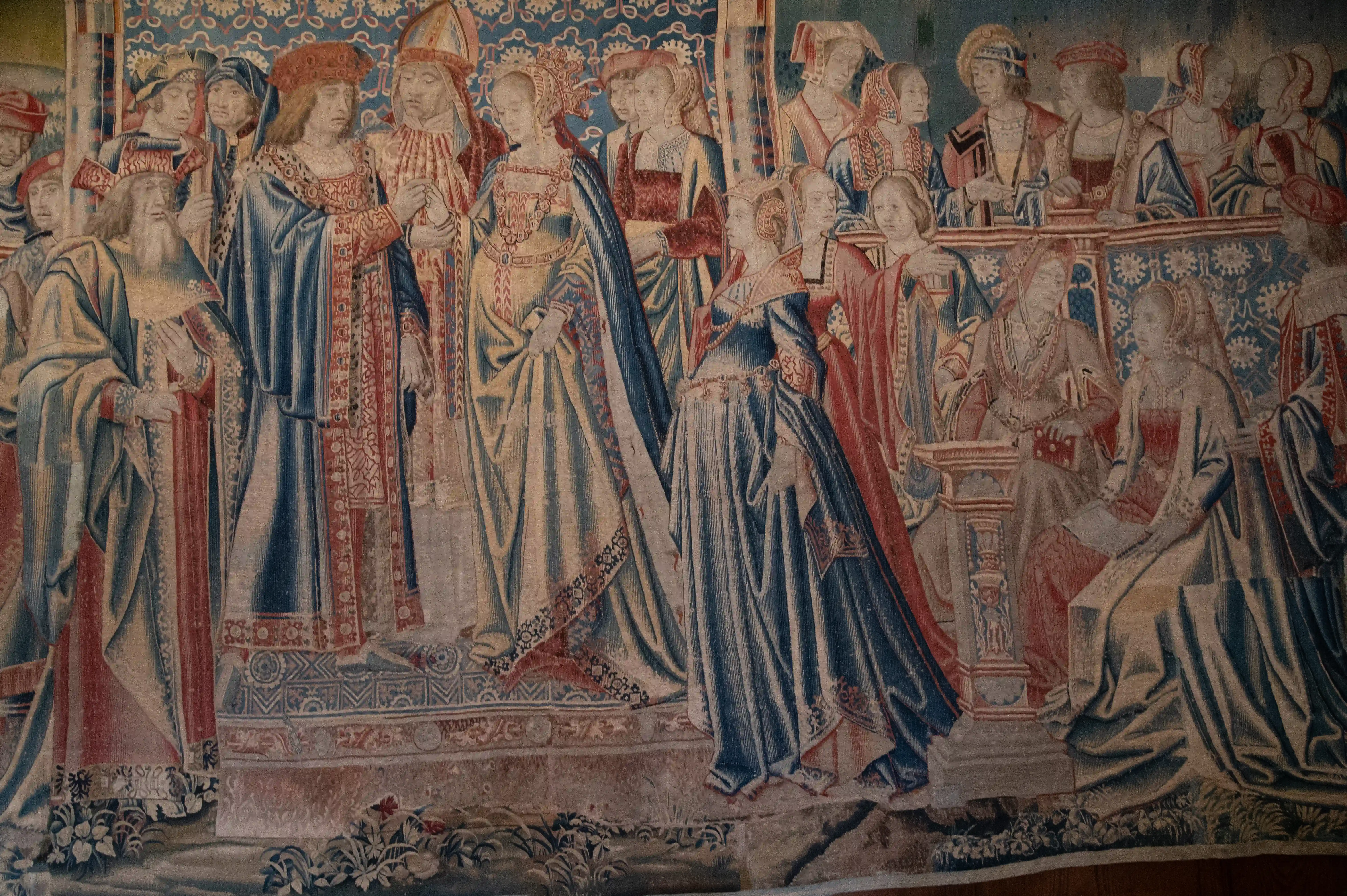 A medieval tapestry showing a group of men and women in elaborate, colourful clothing standing and seated, set against a patterned blue background.