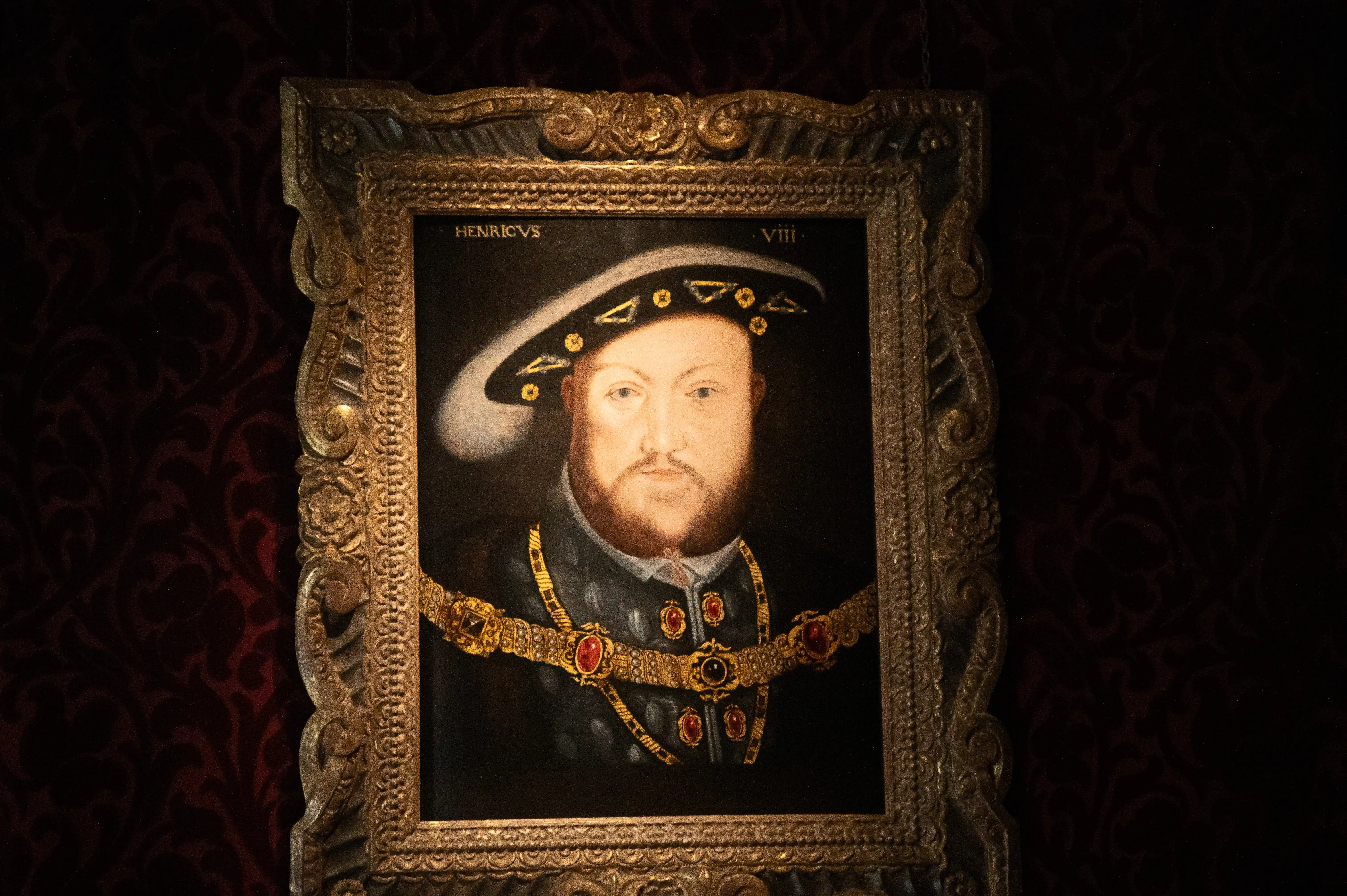 A portrait of King Henry VIII, wearing a hat adorned with jewels and a heavy gold chain. The painting is enclosed in an ornate gilded frame.