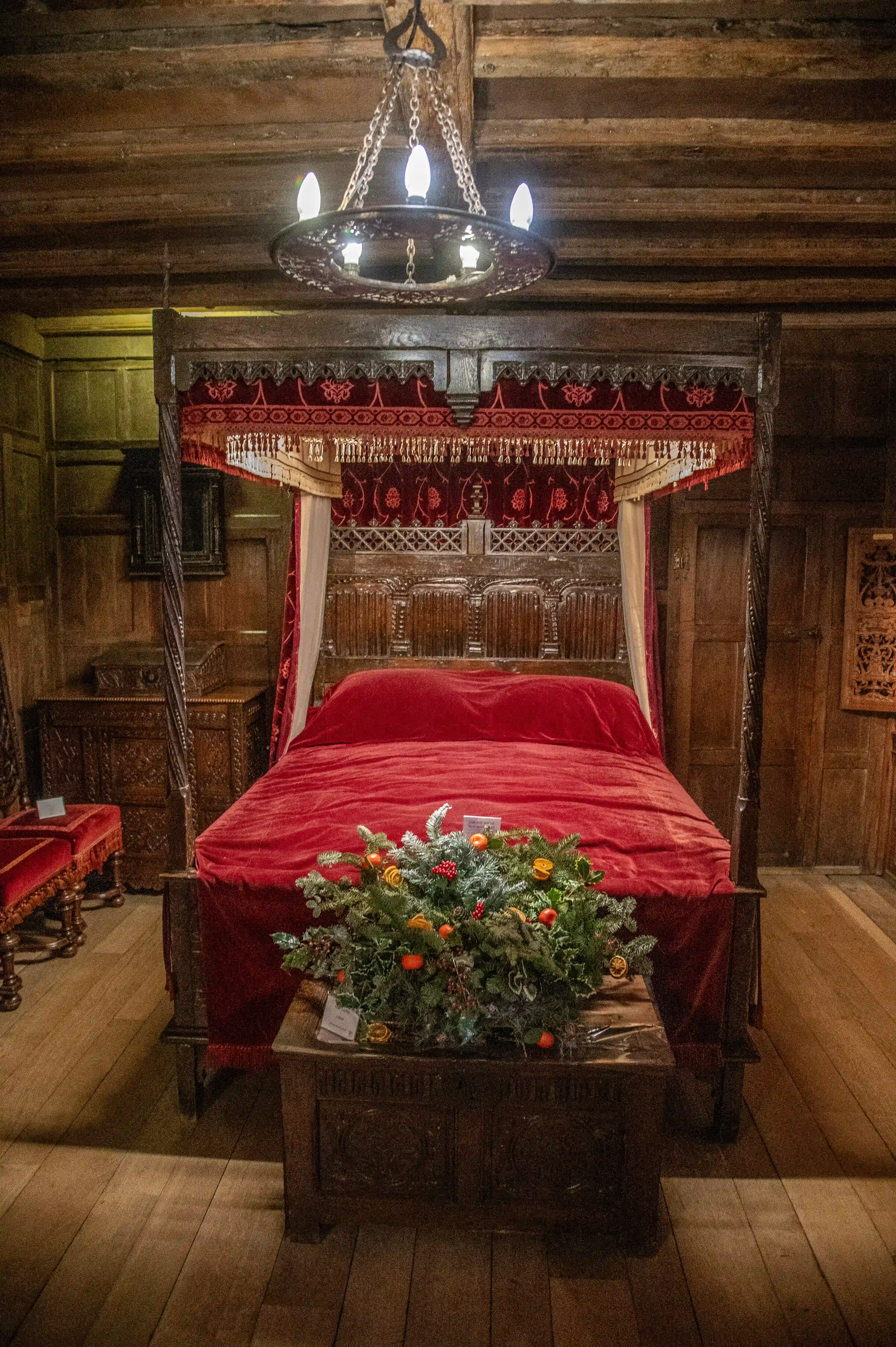 A dimly lit, ornate four-poster bed with red bedding and drapes, featuring a Christmas wreath on a chest at the foot of the bed in a historical room.
