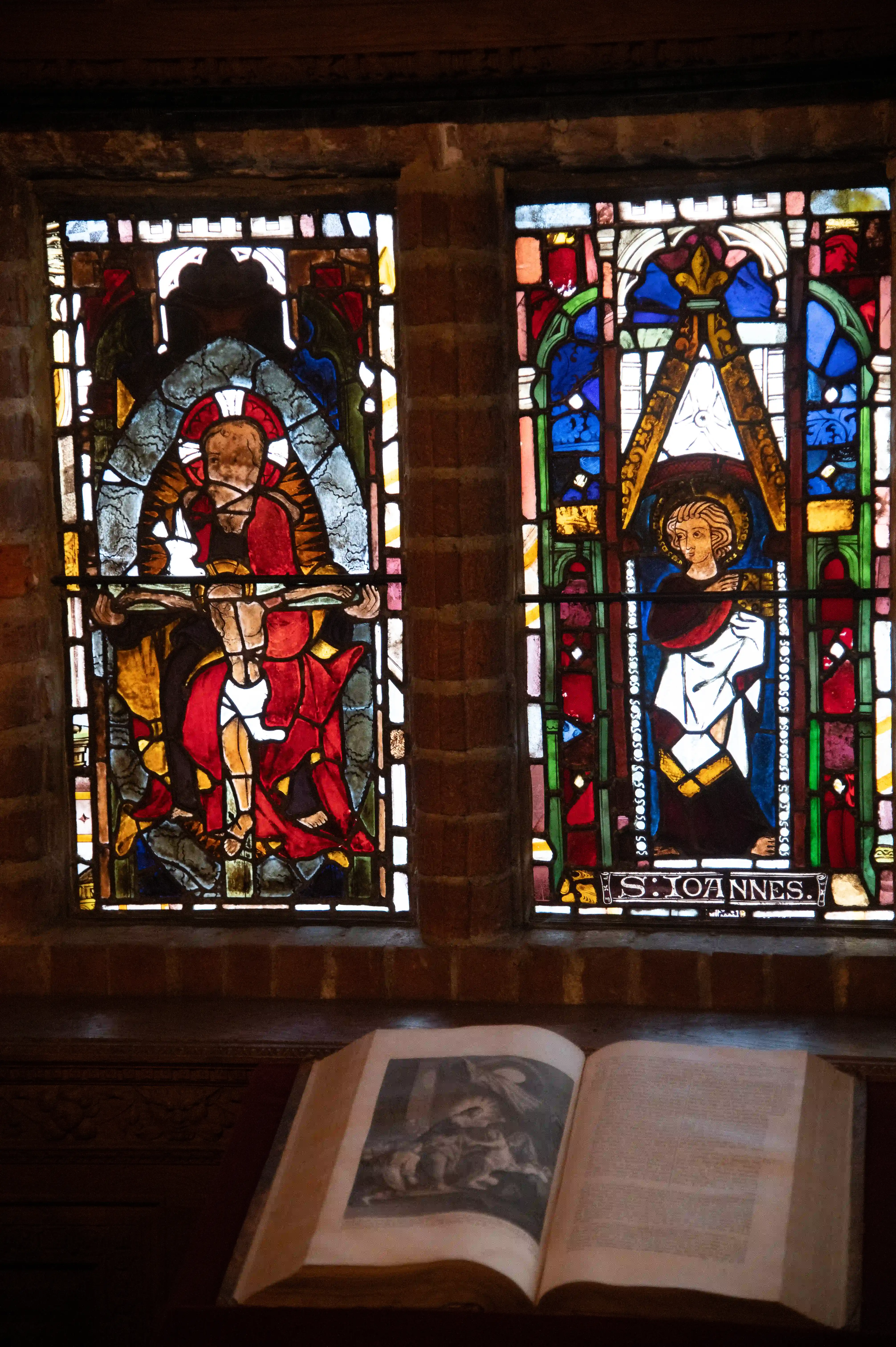 A close-up of two religious stained glass windows in a church. The left window depicts the crucifixion of Jesus Christ. The right window depicts a figure identified as 