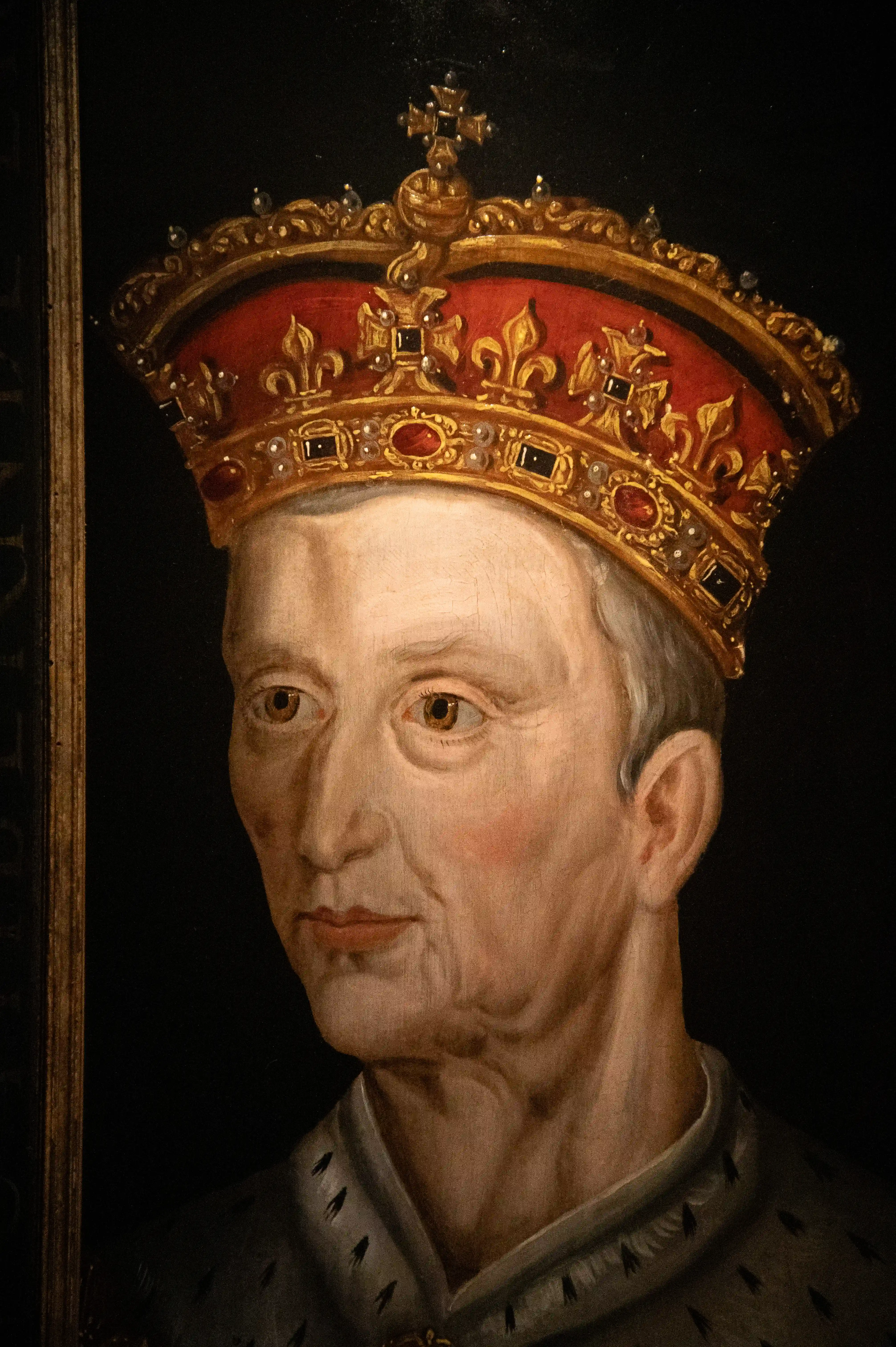 A detailed, close-up portrait of an elderly man wearing an ornate golden crown adorned with red gemstones and fleur-de-lis symbols. He is dressed in a fur-lined robe, suggestive of royalty. The painting style is traditional, with a dark background that accentuates the subject's features and the richness of his regalia. The subject appears to be looking slightly to the left of the viewer, with a solemn expression.