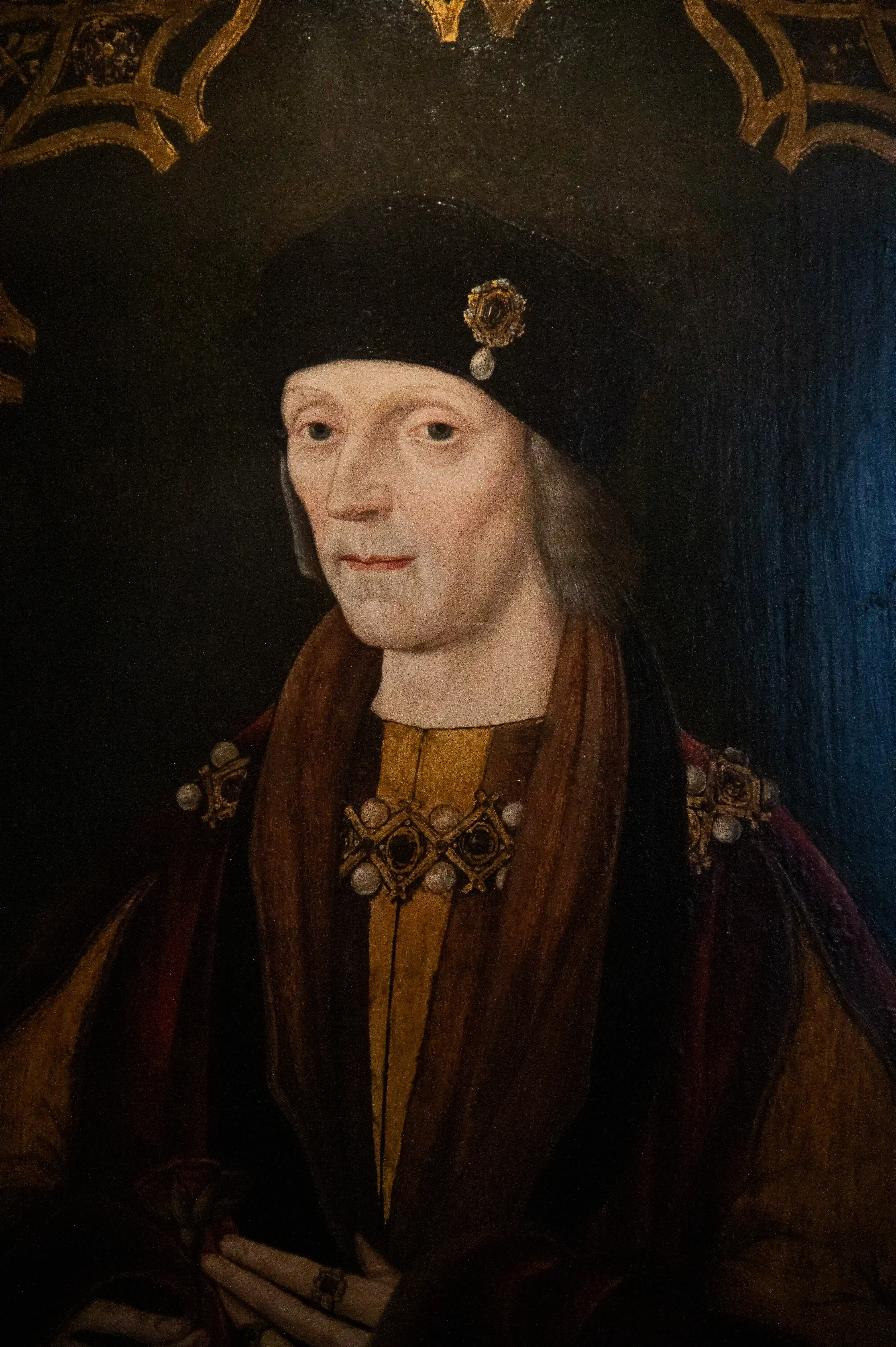 Oil painting portrait of a fair-skinned man wearing a black Tudor hat with a jewelled brooch, a gold tunic, and a red velvet cloak with ornate jewelled trim.