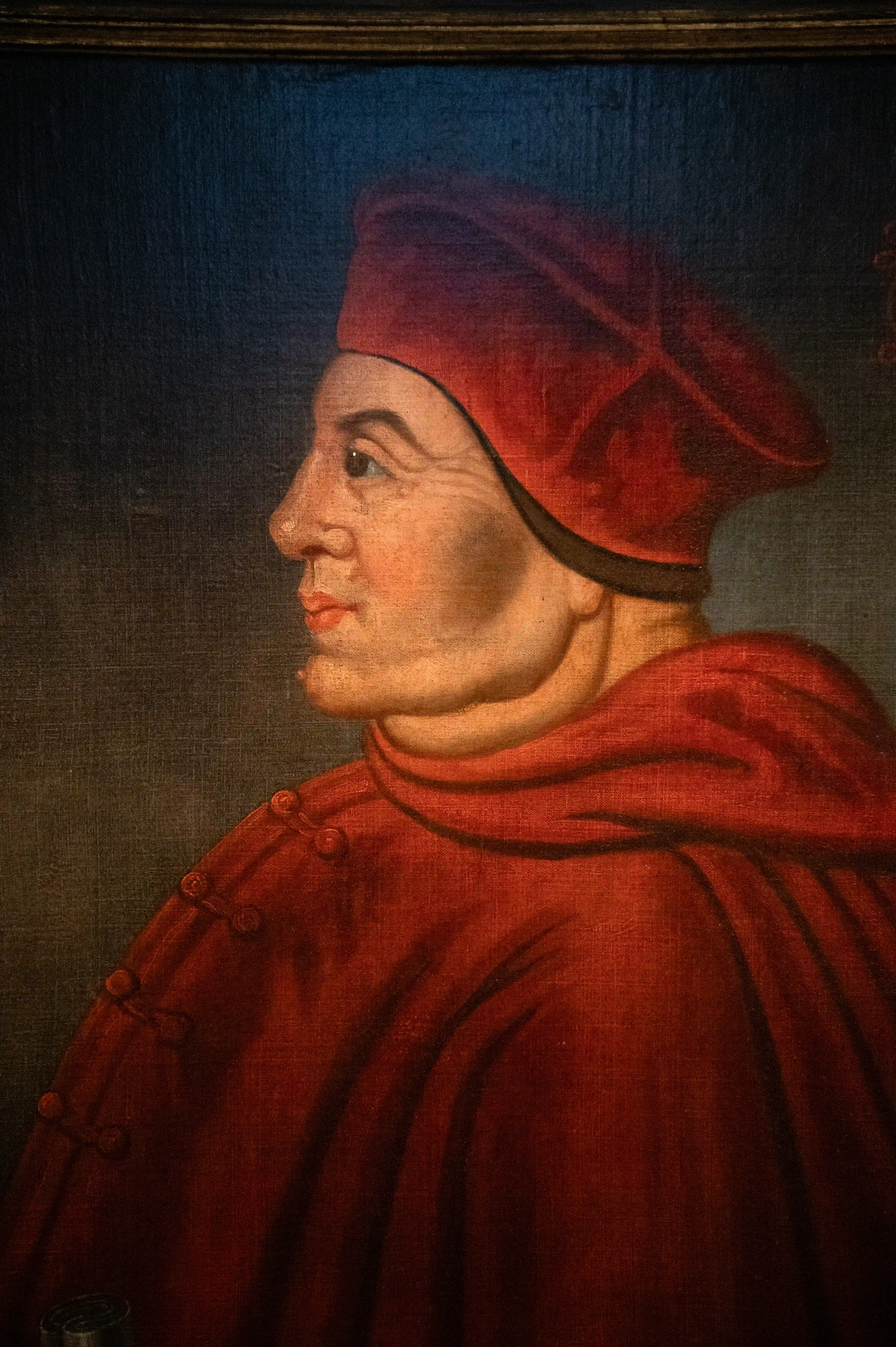 A Renaissance oil painting in profile of a cardinal wearing a red hat and robes against a dark background.