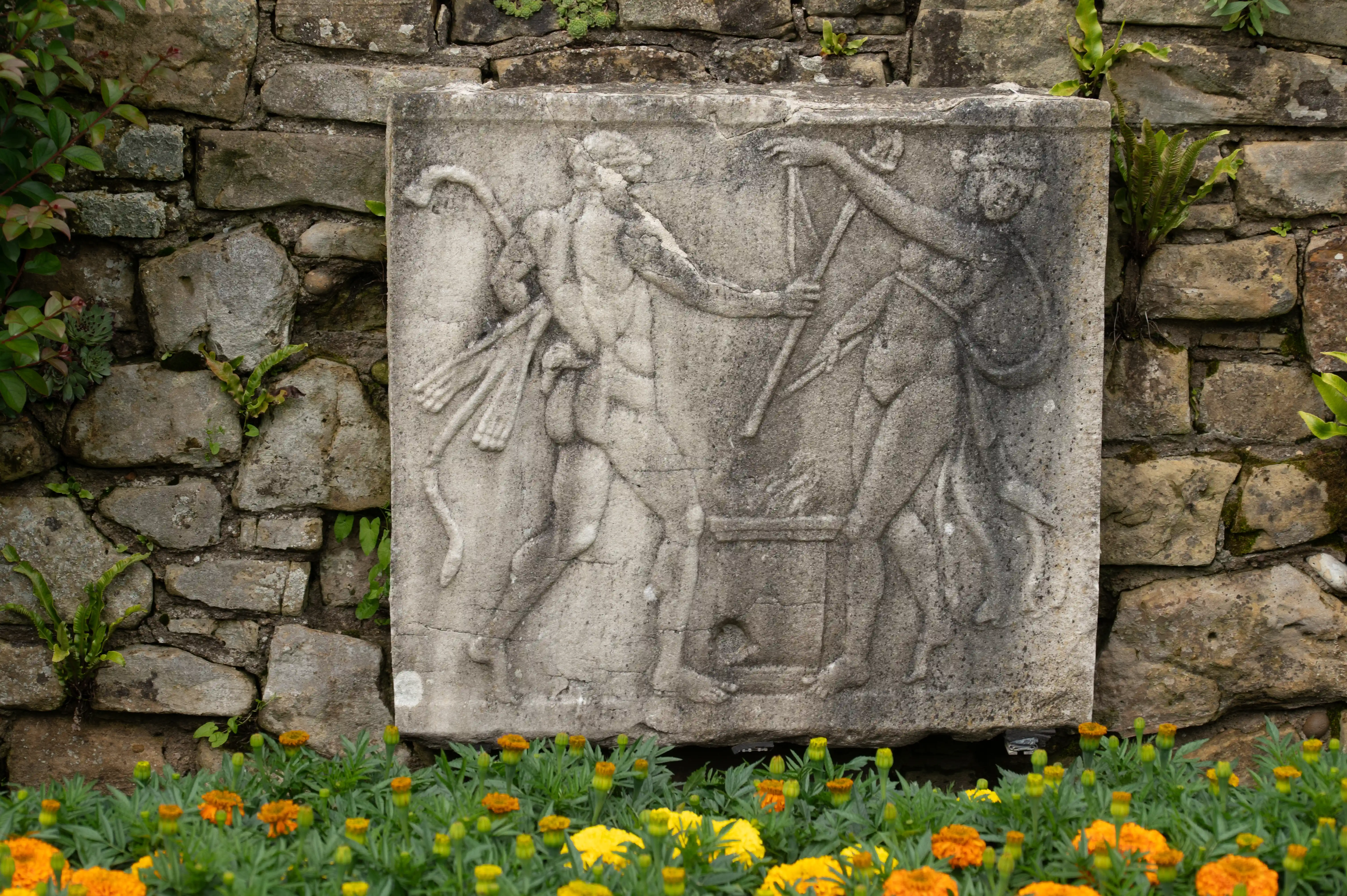 A stone relief carving of two figures in action, set against a textured stone wall, with a border of orange and yellow marigolds in the foreground.