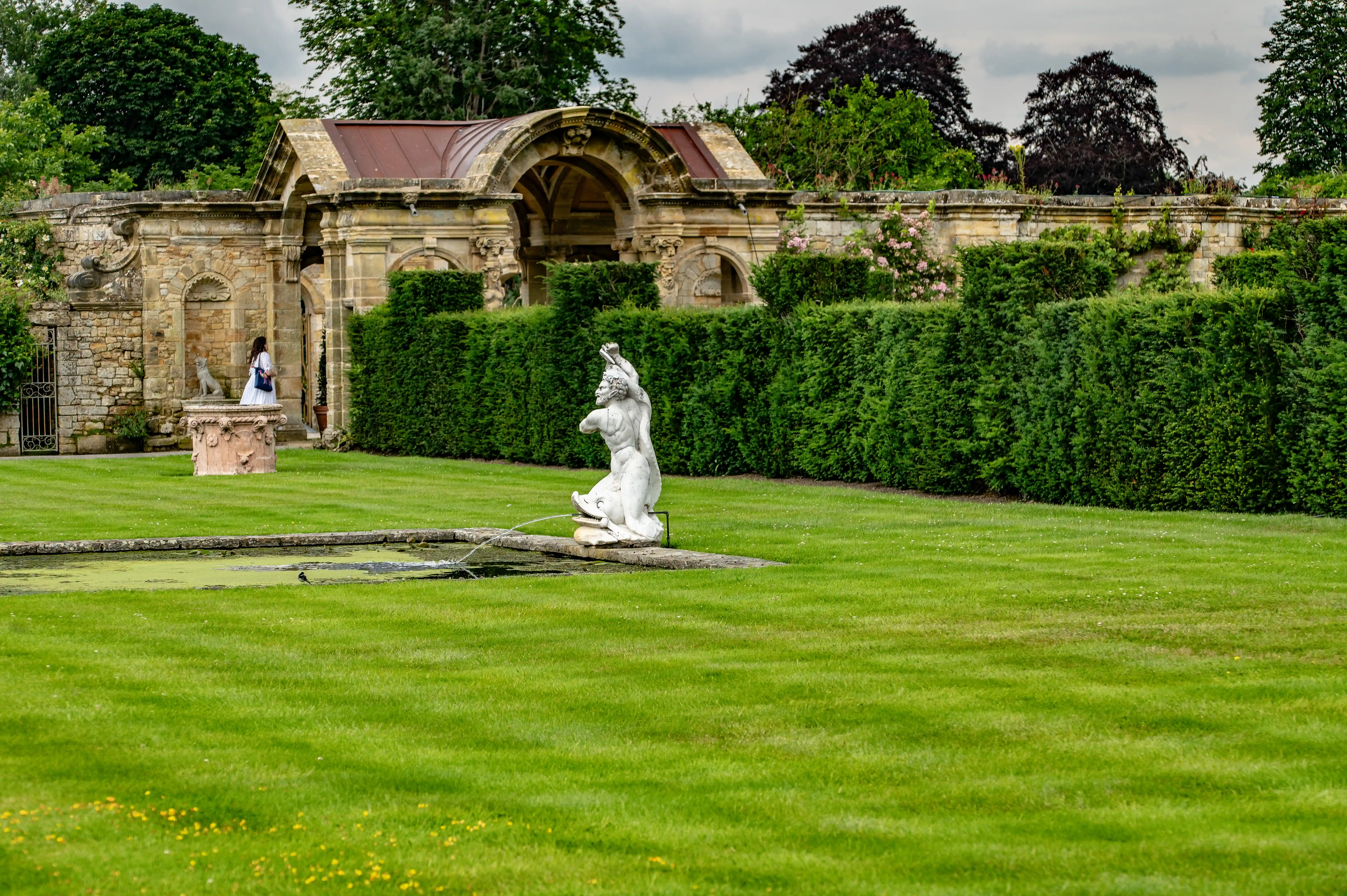 A white marble statue of a man is positioned in a fountain with water spraying. Behind the fountain, a perfectly manicured green hedge runs across the image. To the left of the hedge is a stone wall with arched openings and a person in a white dress is walking. A large stone archway with a copper roof is behind the wall.