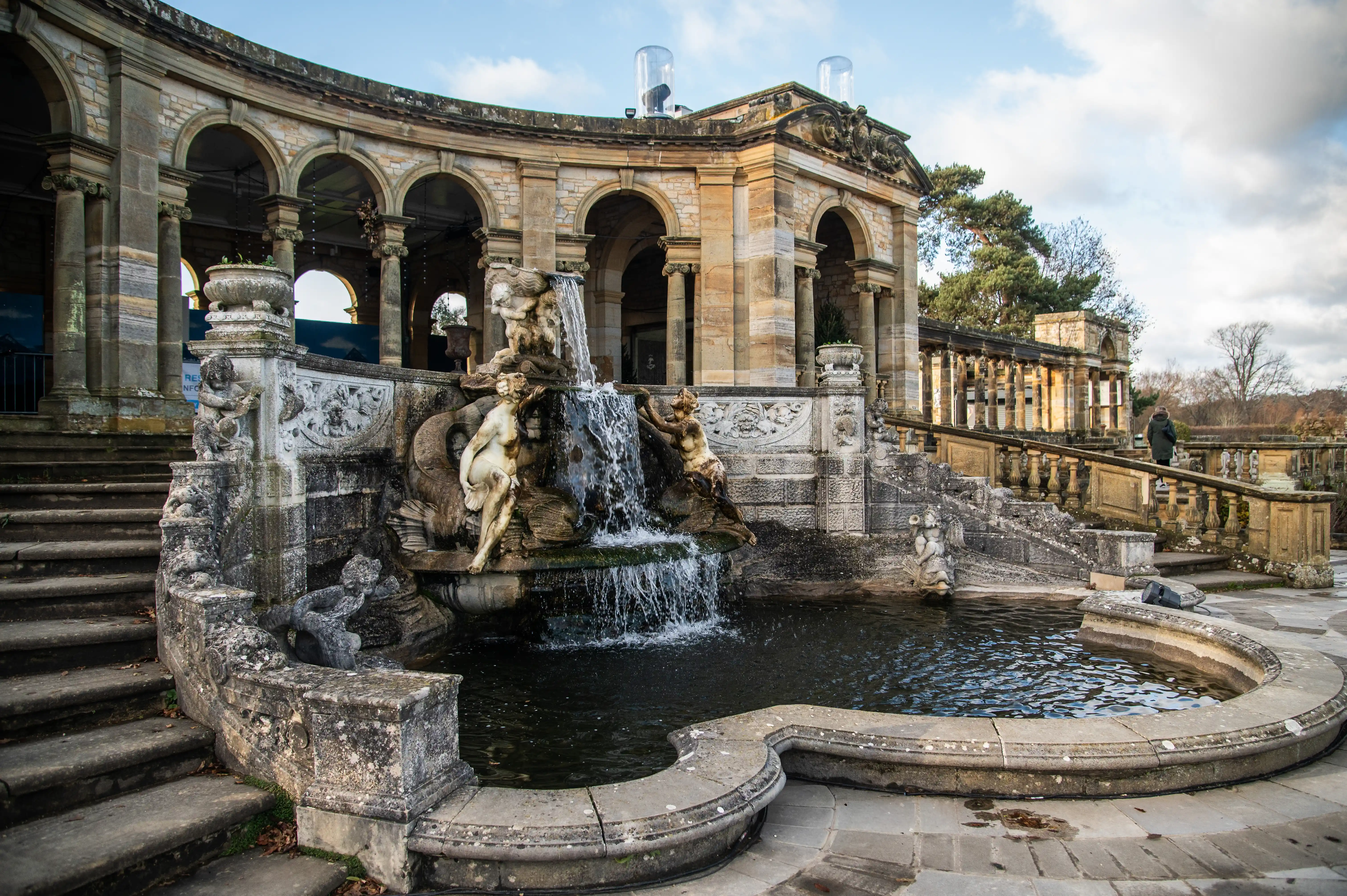 A wide shot of a grand outdoor water feature with classical sculptures. Water cascades down from a central point, flowing into a dark pool. Behind the fountain is a stone building with a curved arcade of arches supported by columns. A person in a green coat is visible walking up a stone staircase to the right.