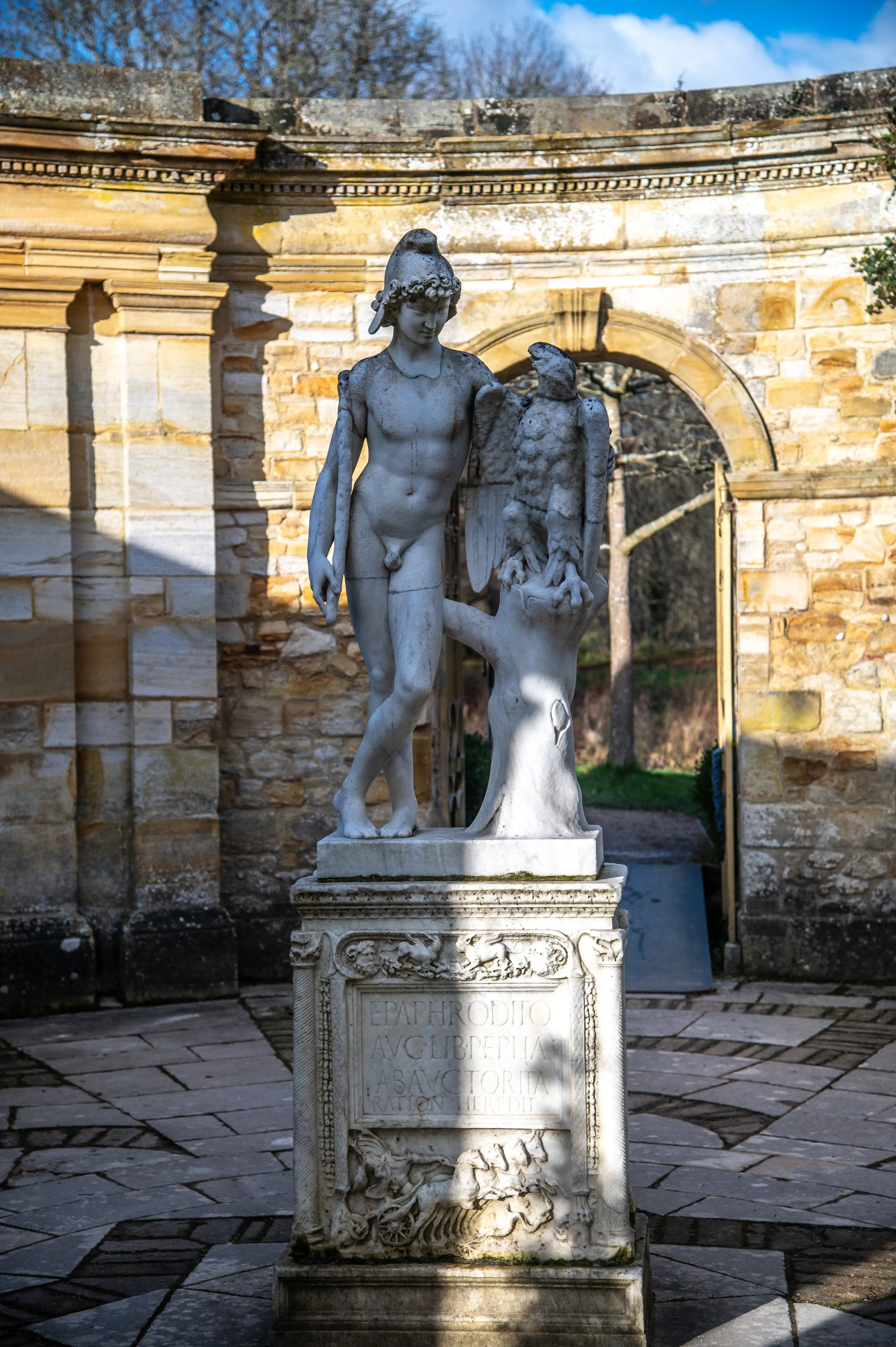 A classical marble statue of a young man with a helmet and a bird of prey perched beside him, standing on an inscribed pedestal in an outdoor courtyard with stone architecture.