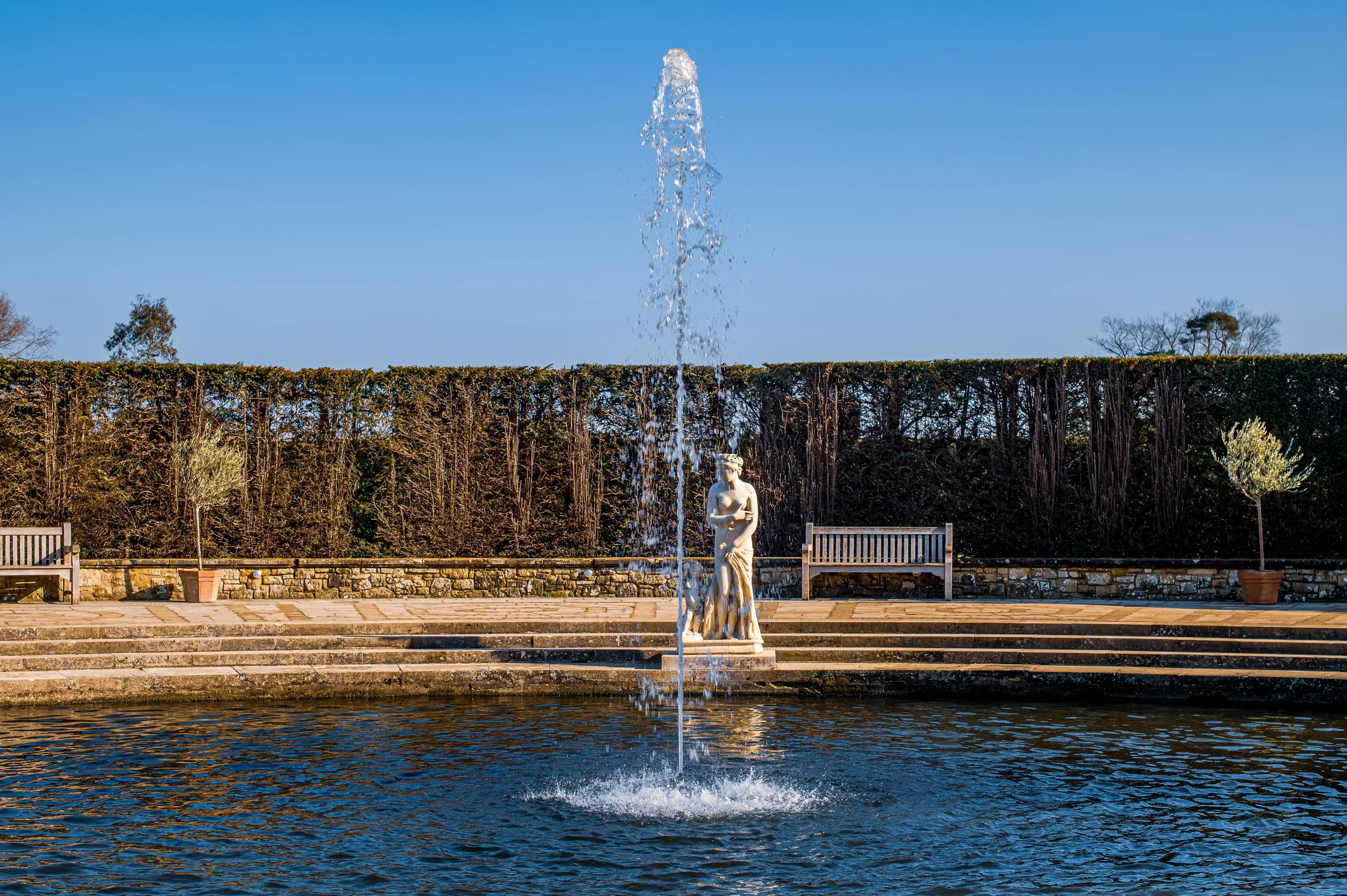 A classical stone statue stands amidst a water fountain in a pond, with terraced steps, stone wall, manicured hedge, and wooden benches in the background under a clear blue sky.