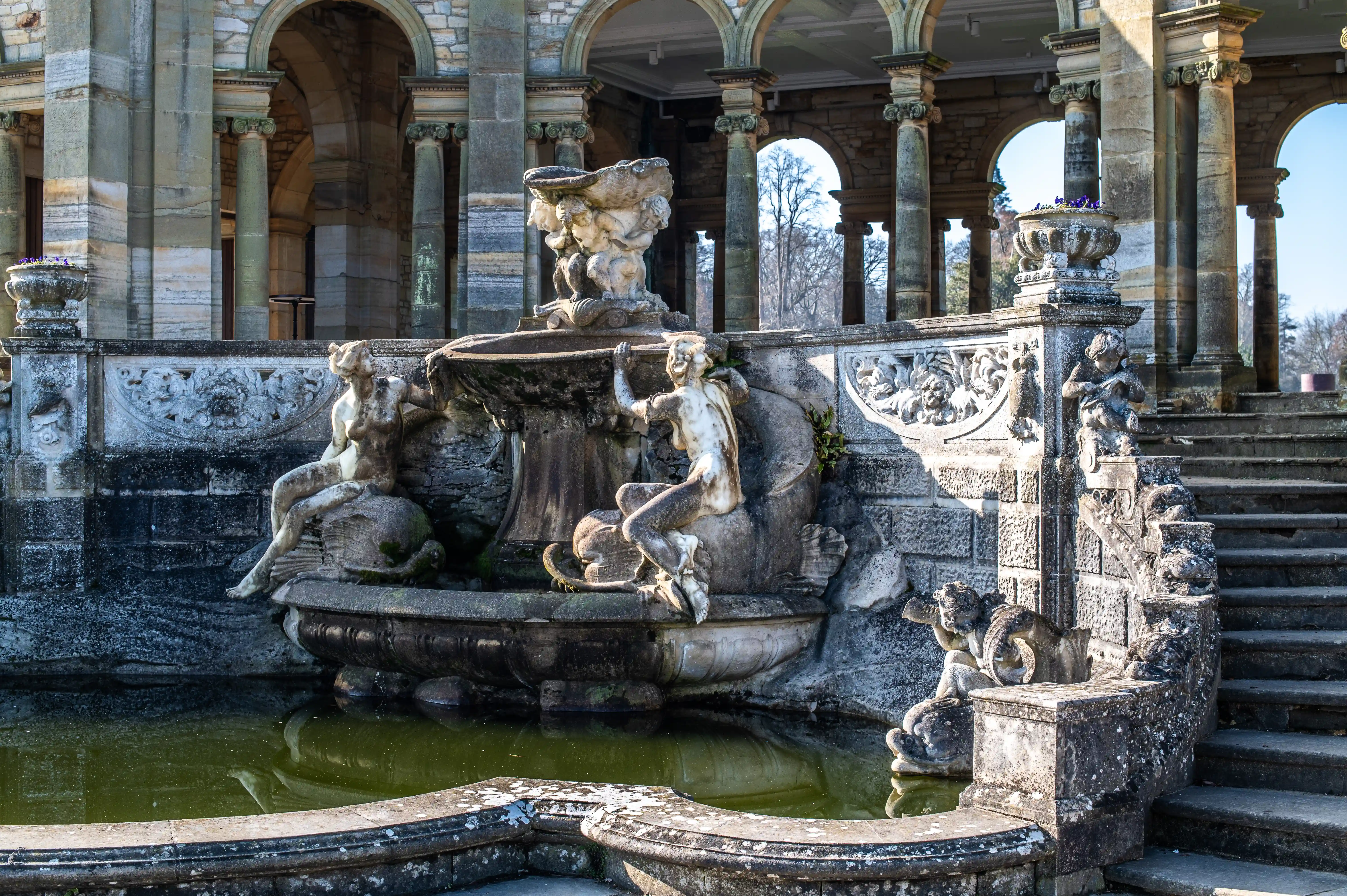 A detailed close-up view of a classical stone fountain featuring sculpted figures and a multi-tiered basin, set against the backdrop of a large, stone building with a series of arches supported by columns. Sunlight illuminates the scene, creating strong contrasts and revealing the aged texture of the stonework.