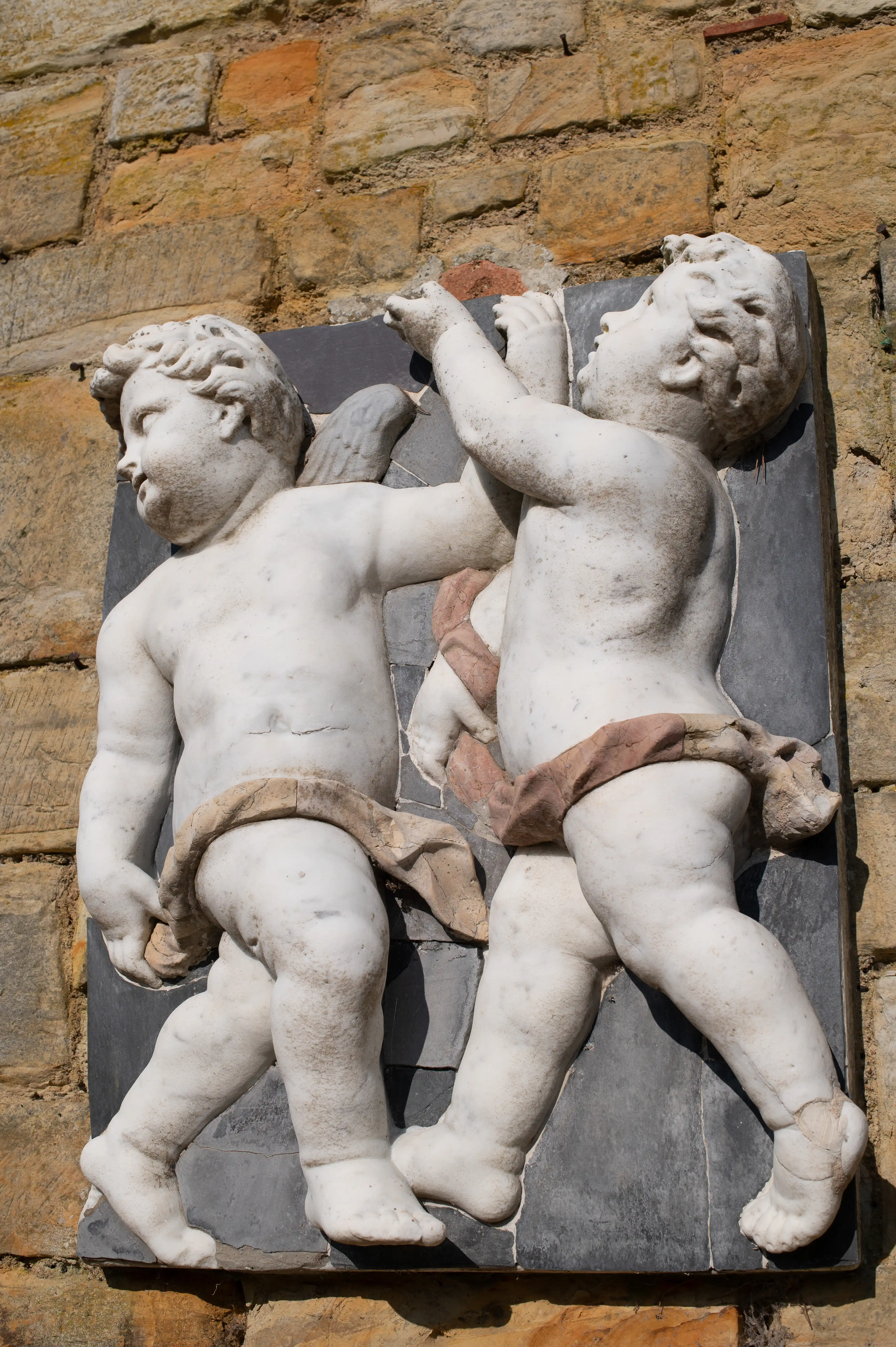 A sculpted marble relief of two chubby cherubic children against a stone wall. One child reaches upwards, the other looks to the left, and both wear drapery around their waists.
