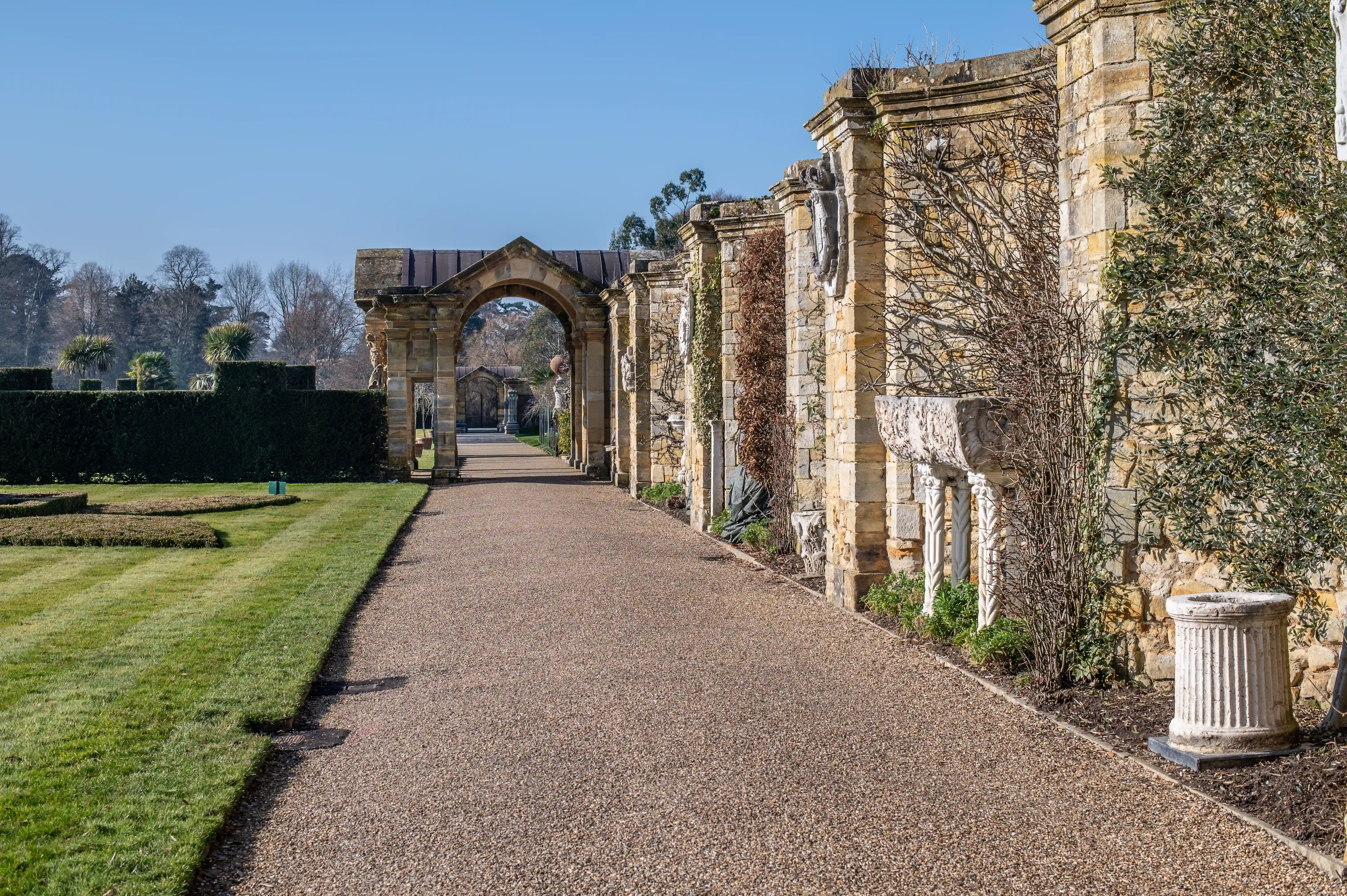 A wide gravel path curves towards an imposing stone archway in a formal garden. To the right, a textured stone wall features decorative sculptures and urns, while to the left, a vibrant green lawn is bordered by neatly trimmed hedges. The sky is a clear, bright blue.